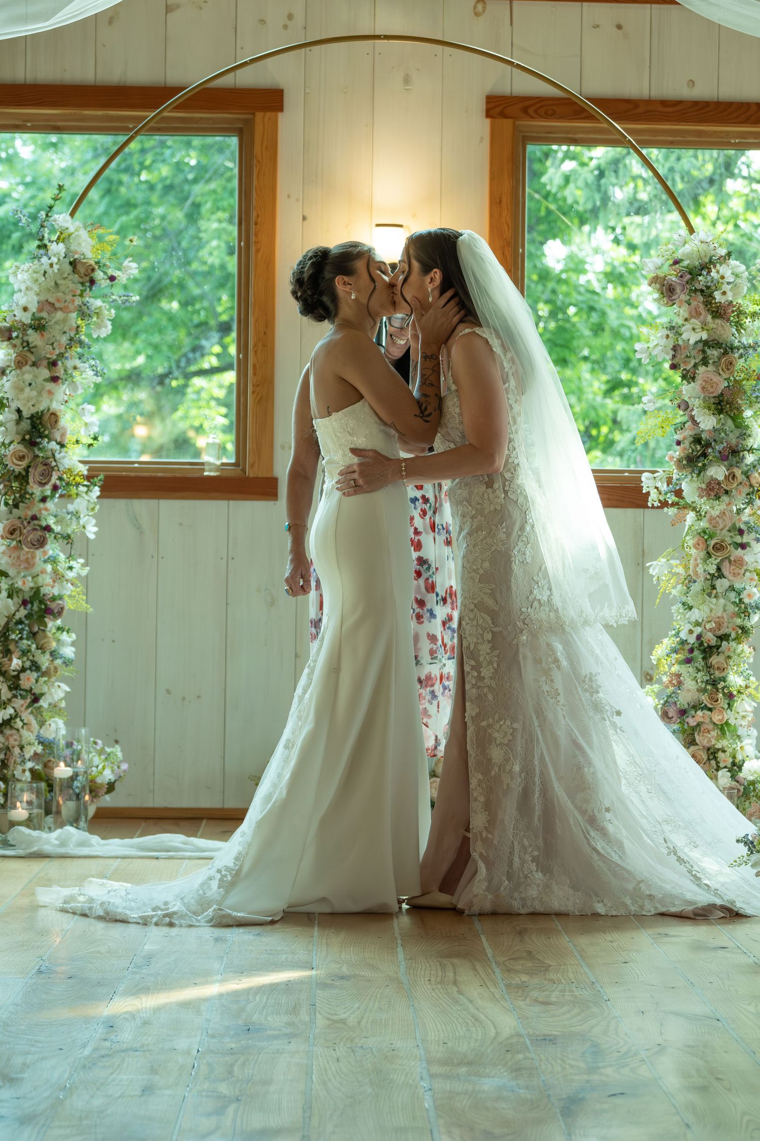 Two brides sharing a kiss under a circular gold arch adorned with white and pink flowers in a bright chapel.