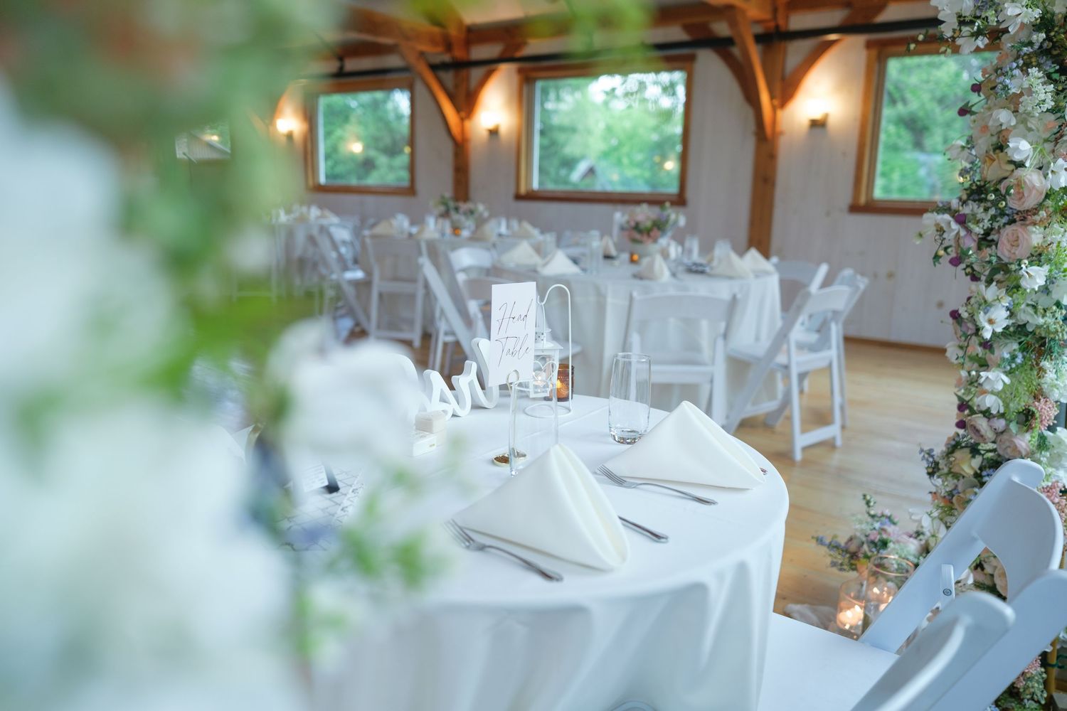 Elegant wedding reception hall with white round tables, folded napkins, floral arrangements, and natural light through large windows.