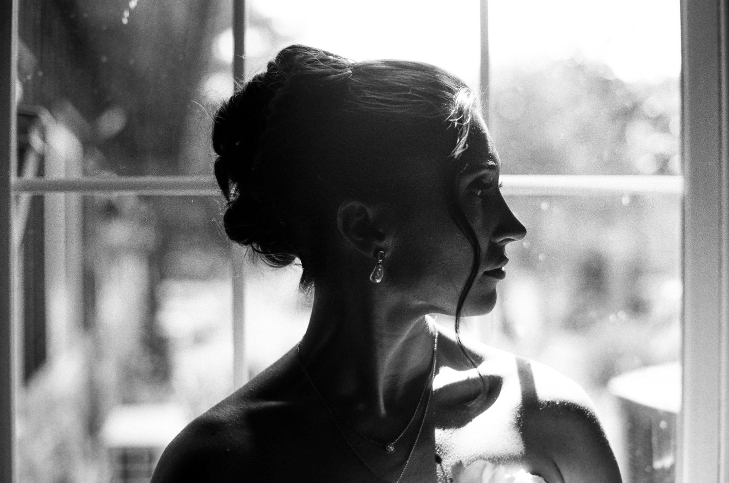 Black and white silhouette portrait of a bride in profile gazing through a window on her wedding day.