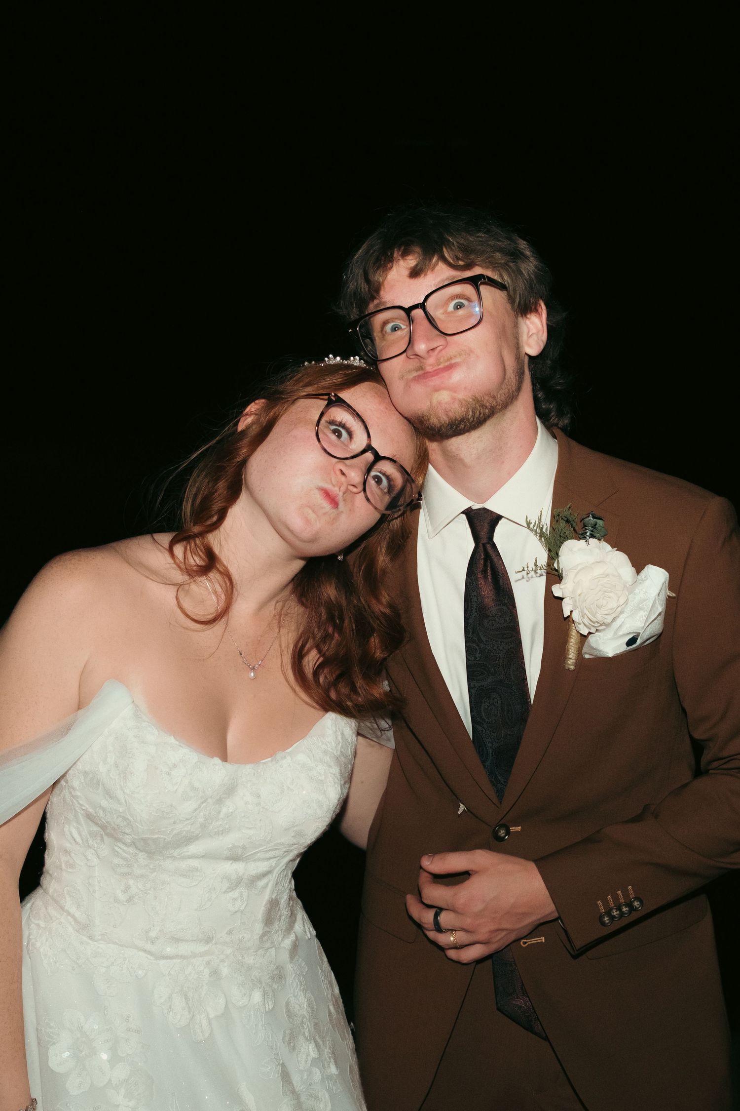 A bride and groom wearing novelty glasses make silly faces at their wedding reception, dressed in white gown and brown suit.