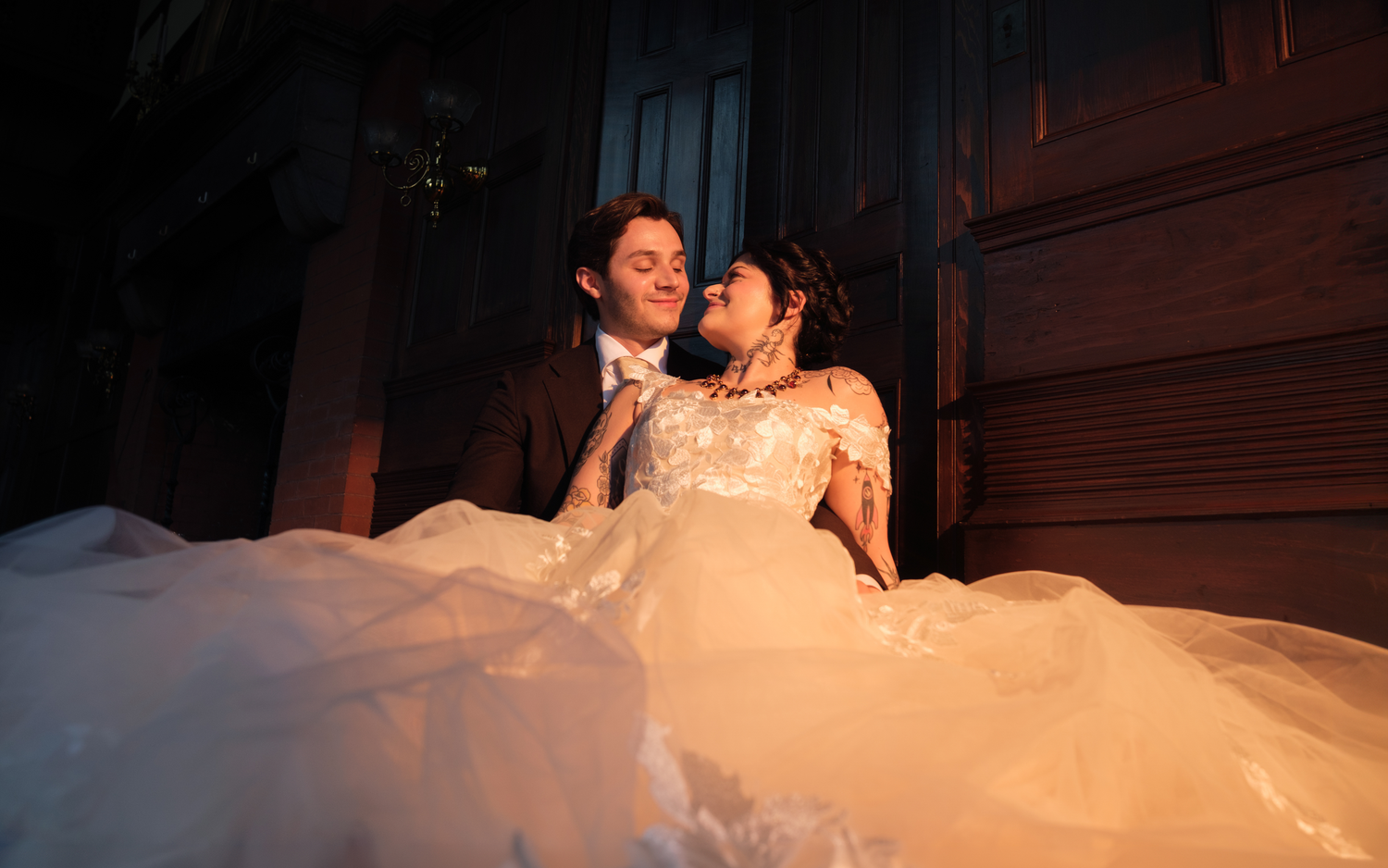 A bride in a white lace gown and groom in a dark suit share a romantic moment in warm golden light.