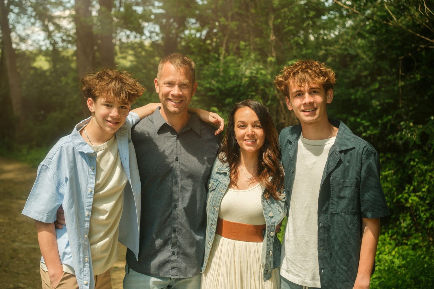 Family of four smiling outdoors in a sunlit forest setting, posing together for a portrait photo.