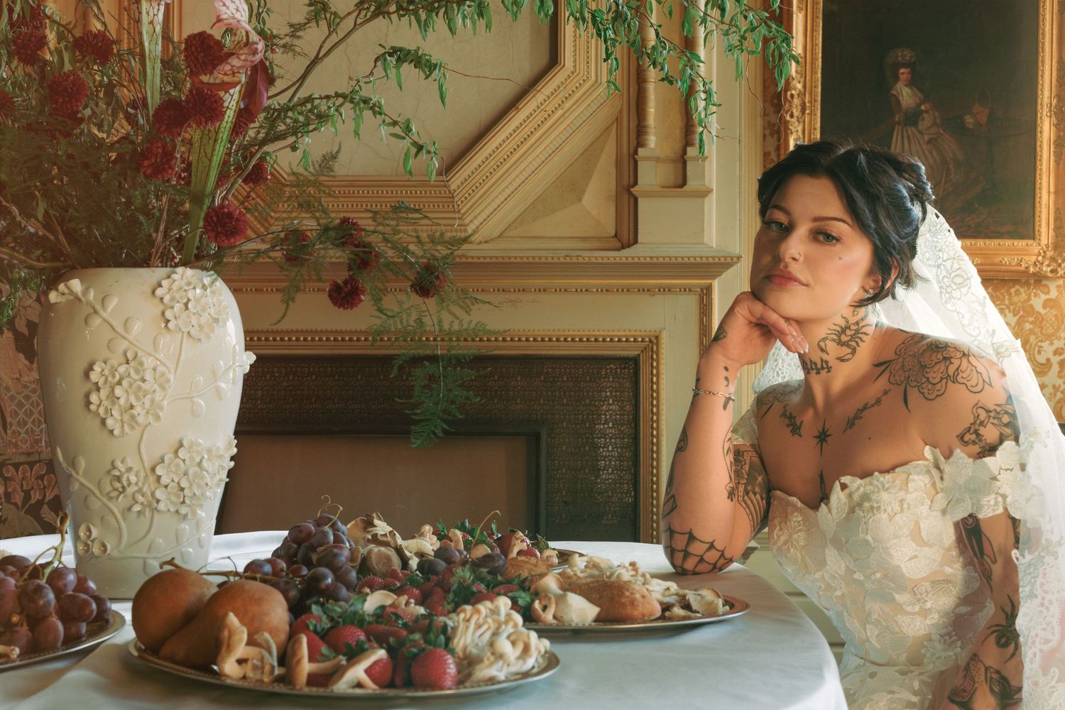 Tattooed bride in lace wedding dress and veil sits at elegant table with charcuterie board and ornate vase.