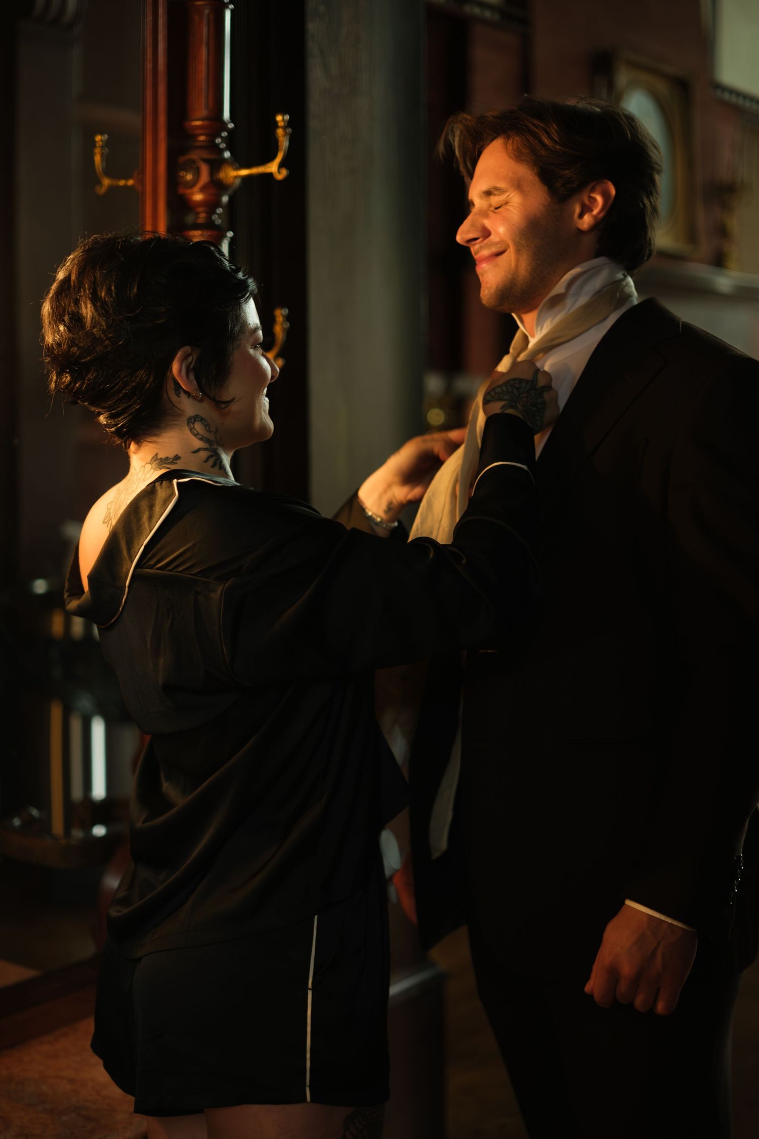 A woman adjusts a man's cravat as he smiles in a dimly lit, elegantly dressed period setting.