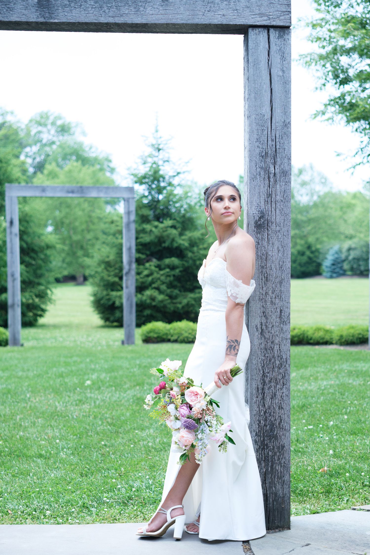 Elegant bride in off-shoulder white gown holds colorful bouquet while leaning against wooden post in garden.