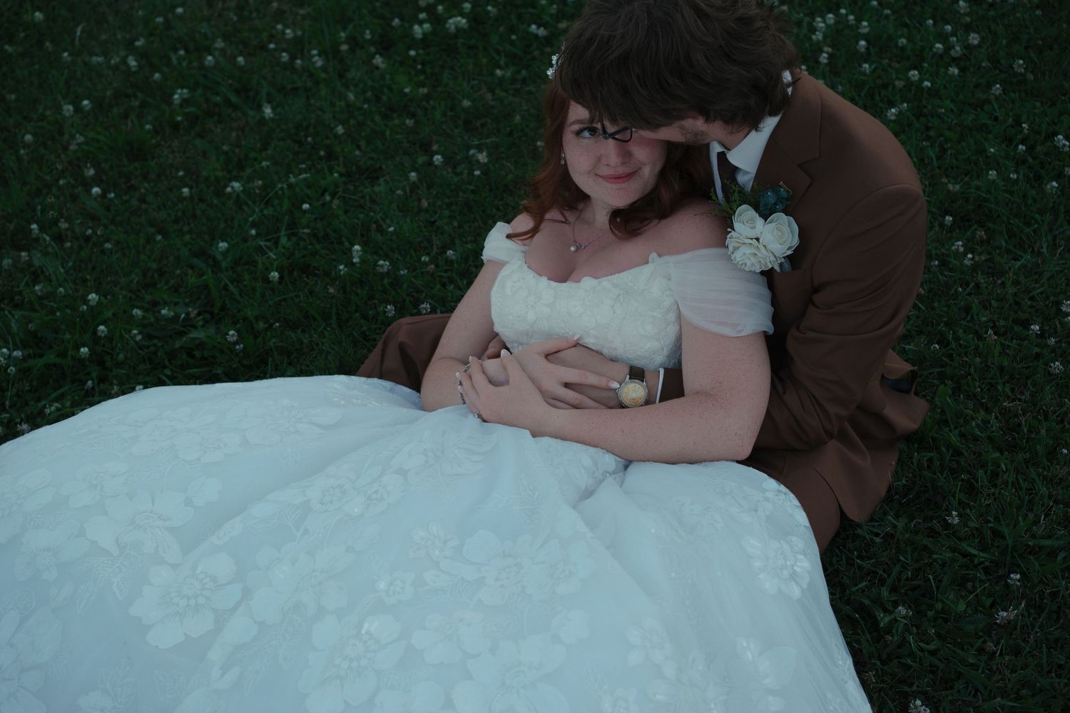 Bride and groom sitting on grass, groom in brown suit embracing bride in white lace gown.