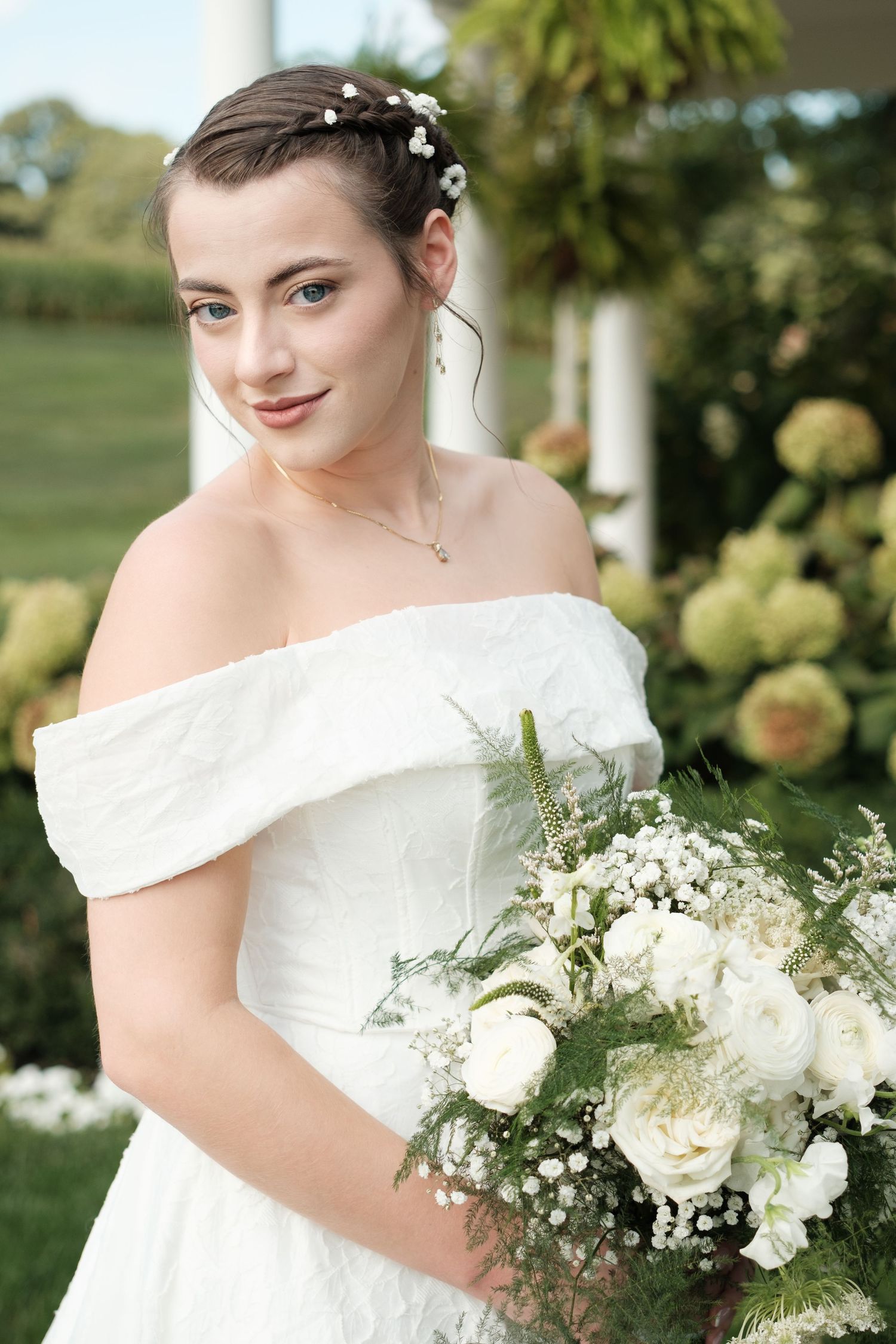 Bride in an off-the-shoulder white wedding gown with a floral hair accessory holds a white rose and baby's breath bouquet in a sunny garden.