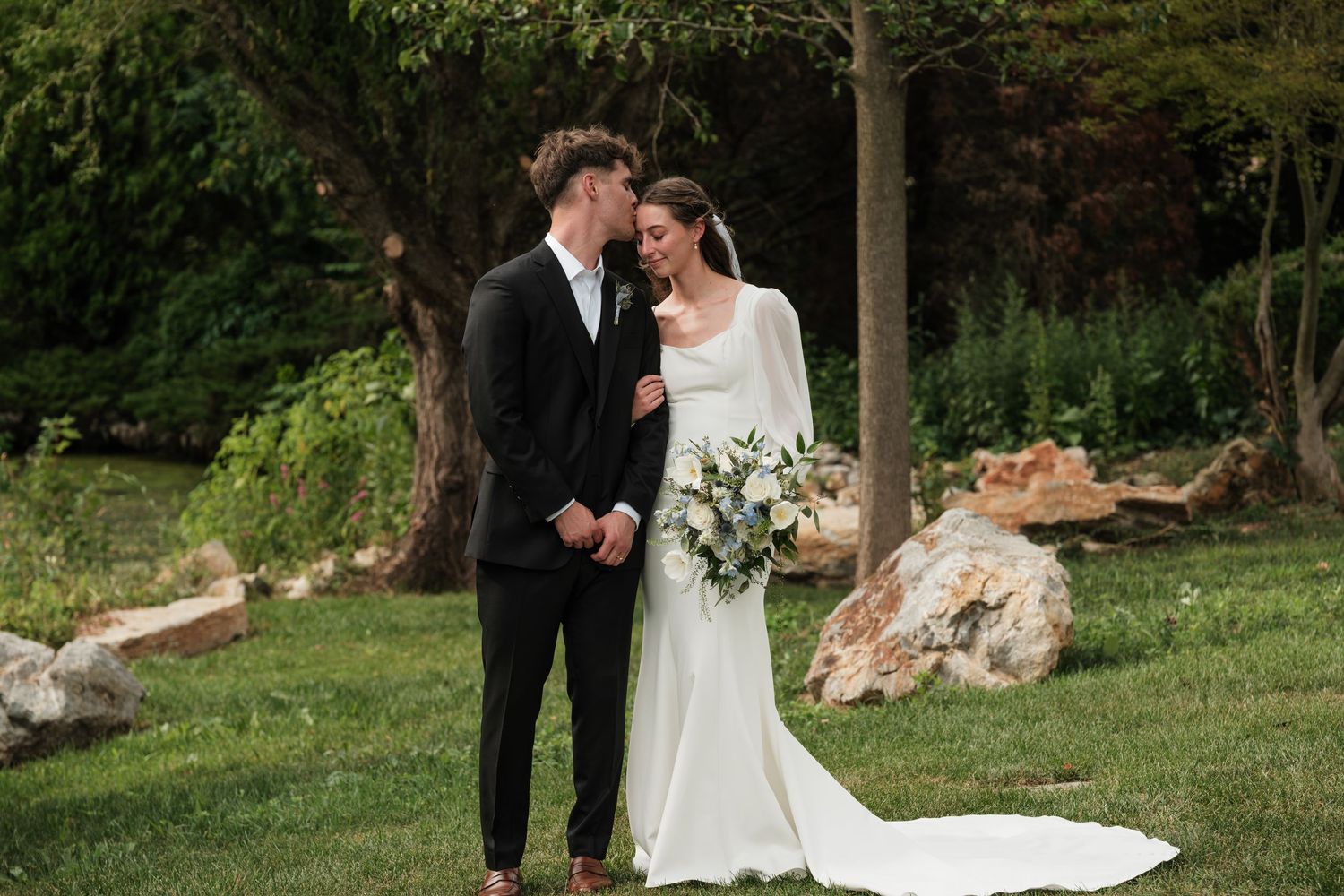 A bride and groom share a tender moment outdoors, she holds a white bouquet in an elegant long-sleeve gown.