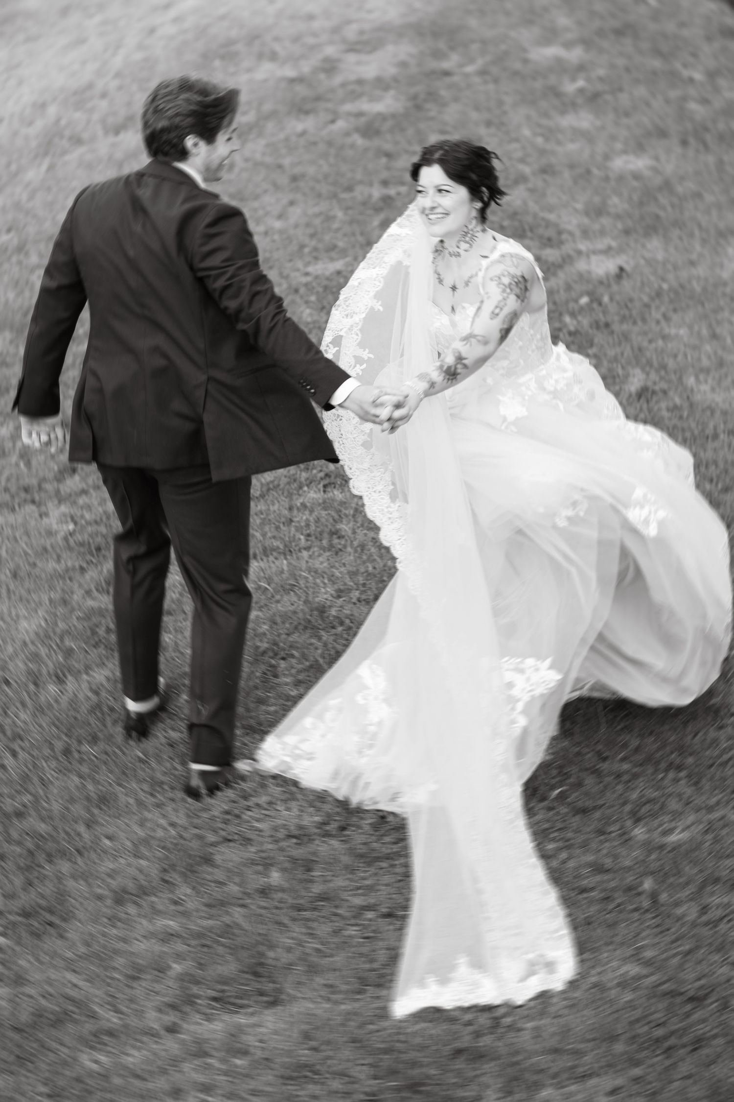 Black and white photo of a bride and groom holding hands, running through a grassy field, her lace gown flowing.