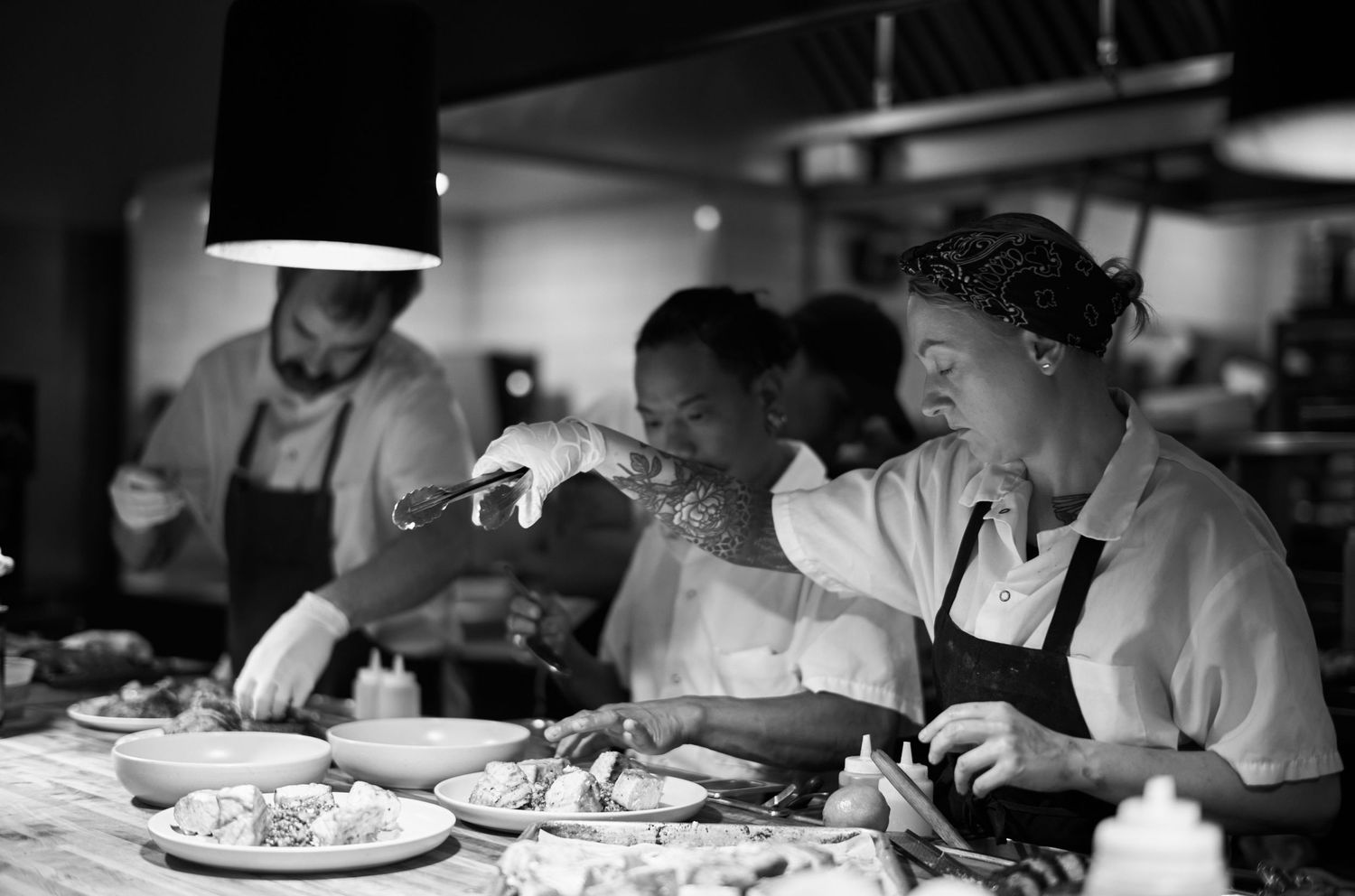 Chefs in white uniforms work diligently in a professional kitchen preparing food in black and white photograph.