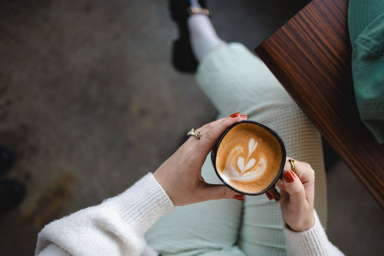 A woman's hands holding a coffee cup with latte art, sitting crossed legged with mint pants.