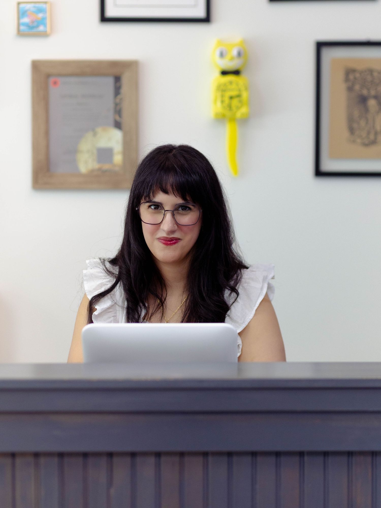 A woman with dark hair and bangs in a retail store sitting behind a till on the computer looking at the camera while working.