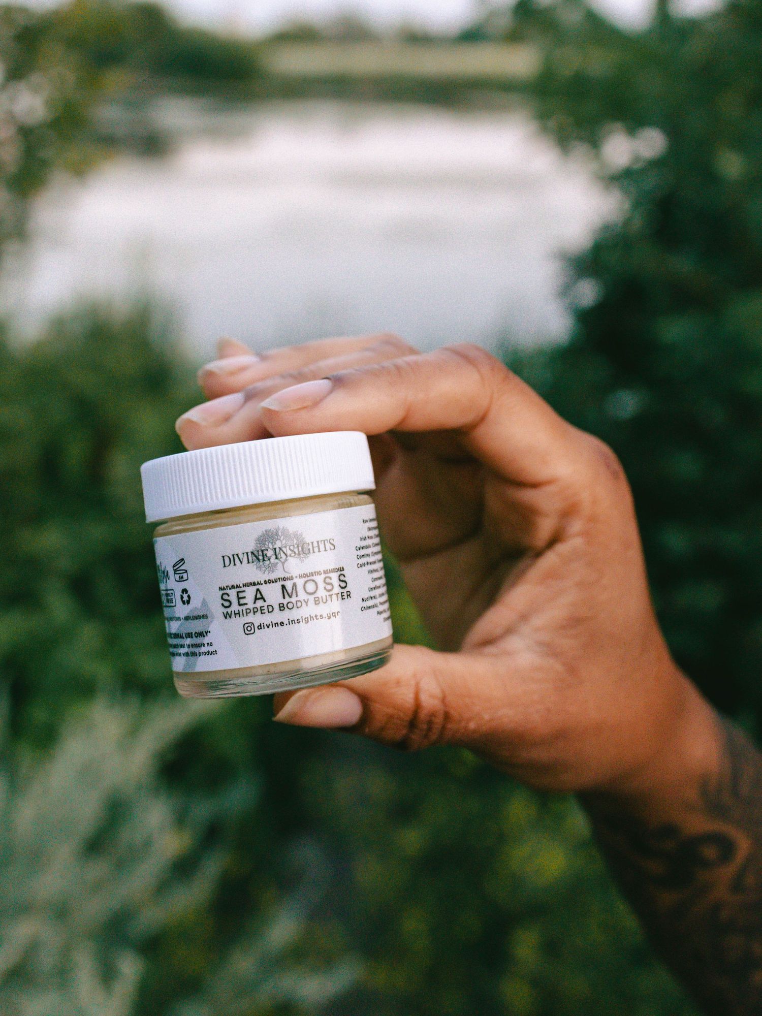 Woman's hand holding skincare product with nature landscape and water in background.