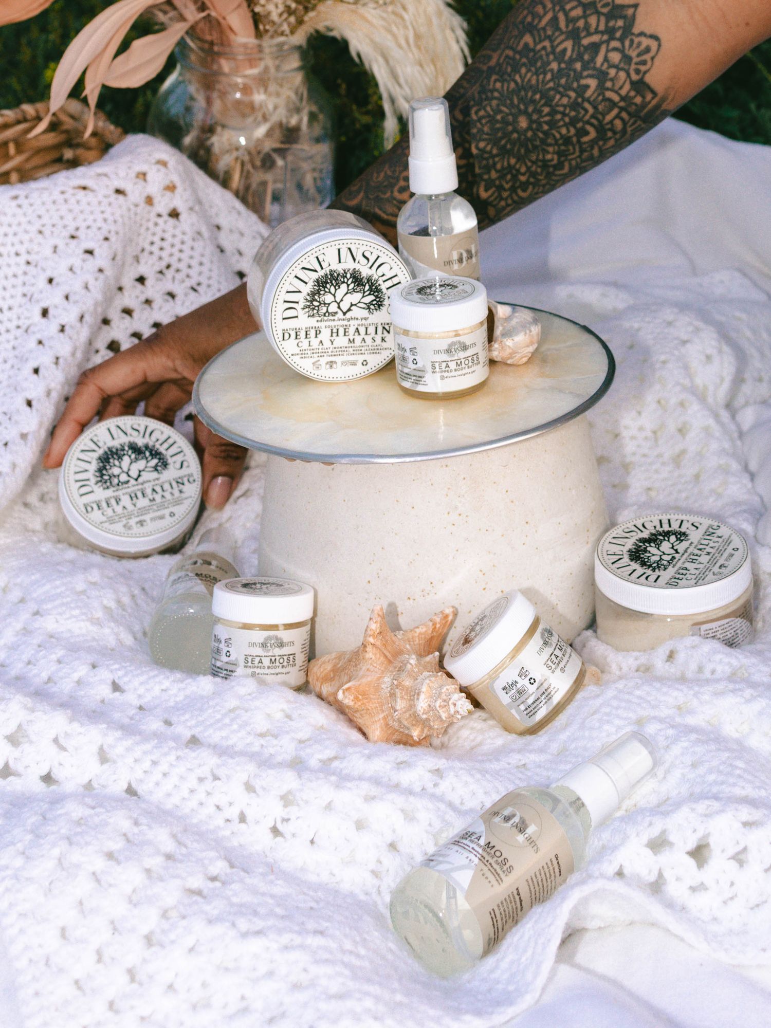 Many skincare products sitting on pearl-like stand, sat on top of white, soft and textured fabrics with woman's arm grabbing product in back