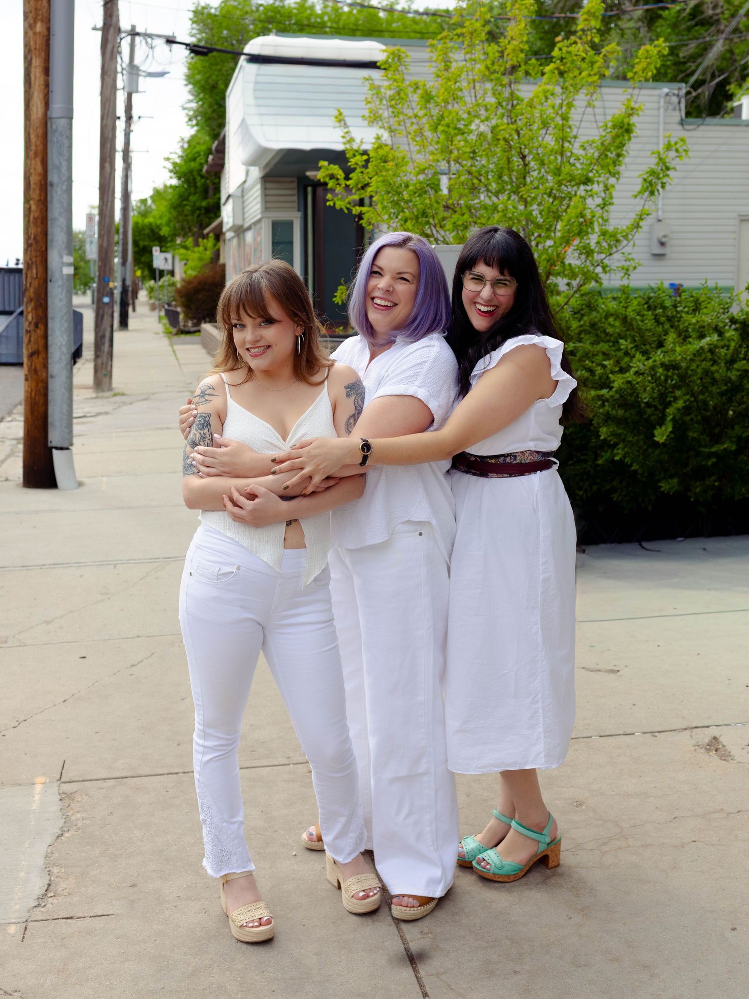 Three woman hugging each other from the side and laughing, all wearing white outfits and posing on a sidewalk outside a retail business.