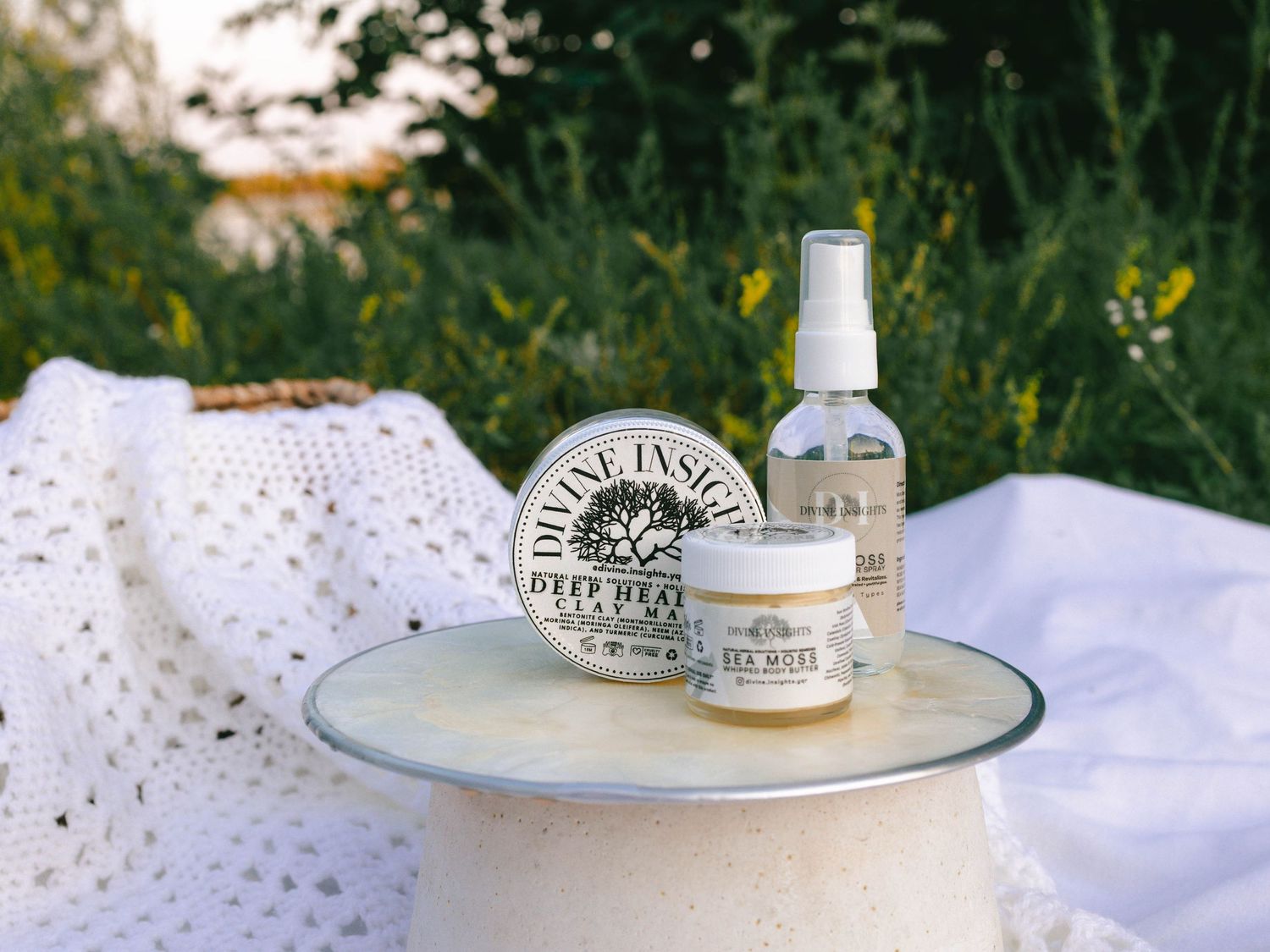 Three skincare products sitting on pearl-like stand, with soft, white textured fabrics behind, with a nature landscape in the background.
