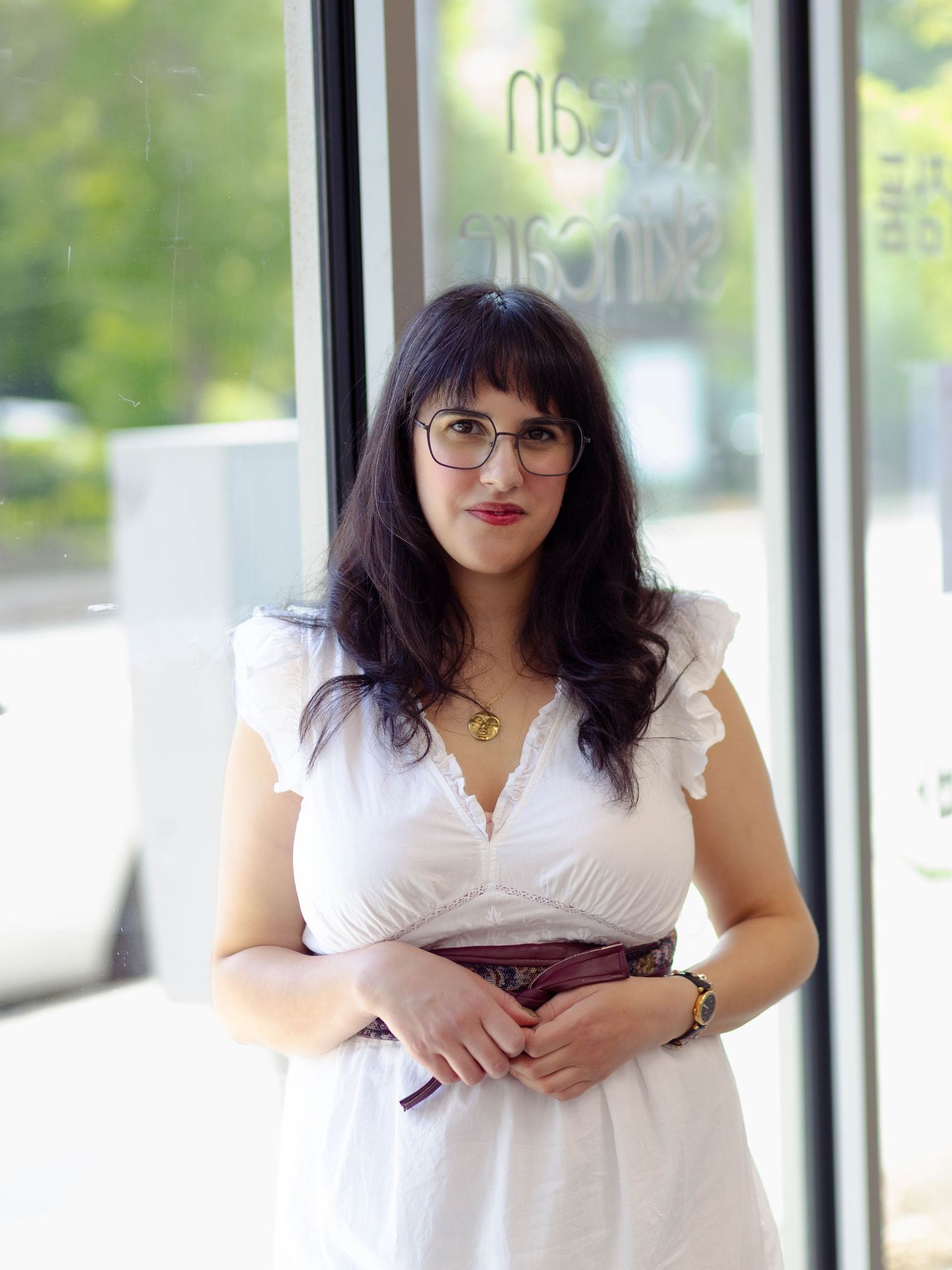 Portrait of a woman with dark hair and bangs, wearing a white dress with brown belt and holding her hands loosely in front of her.
