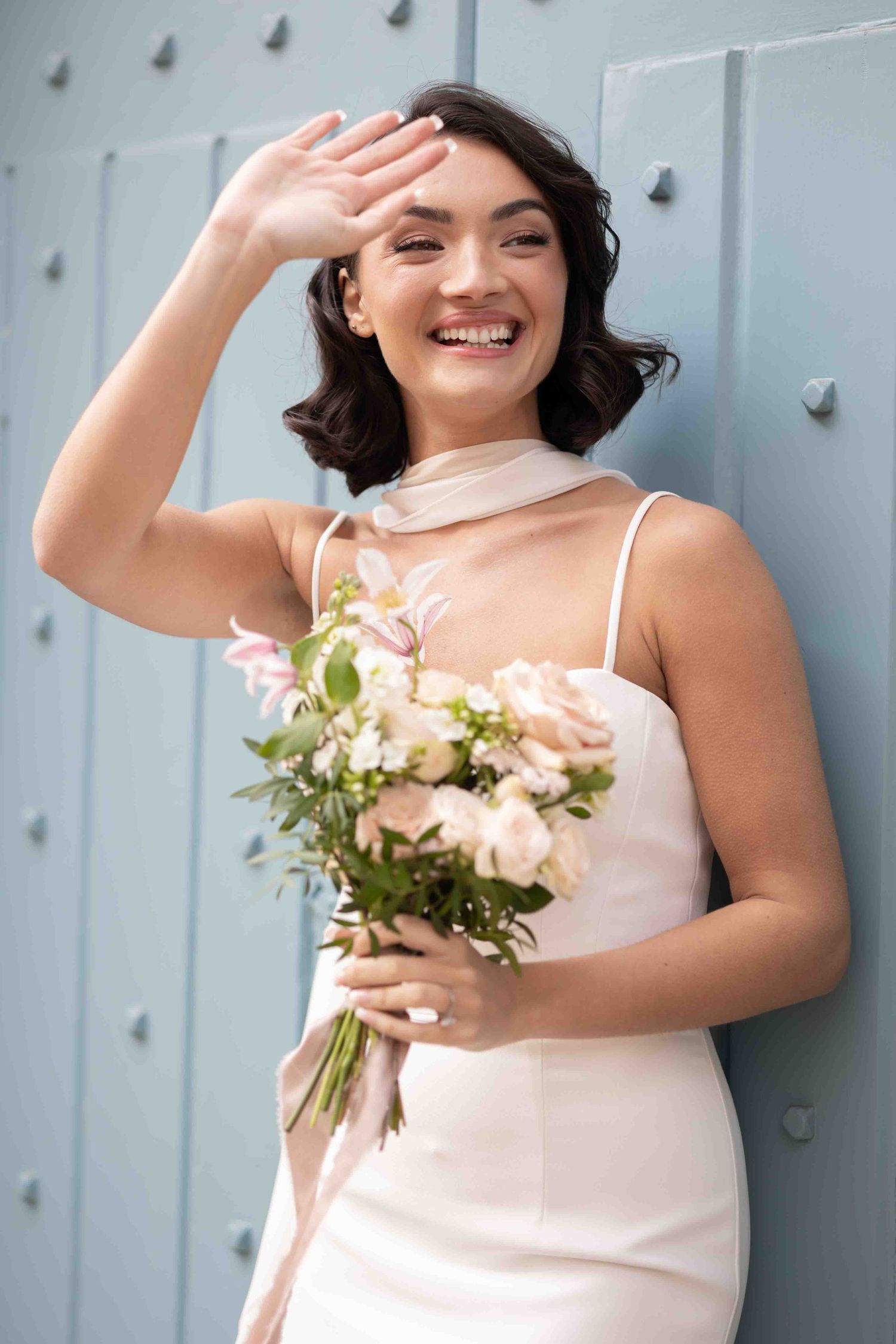 Joyful bride holding a bouquet of blush and cream flowers against a soft blue backdrop in London — timeless fine art wedding portrait by Alb