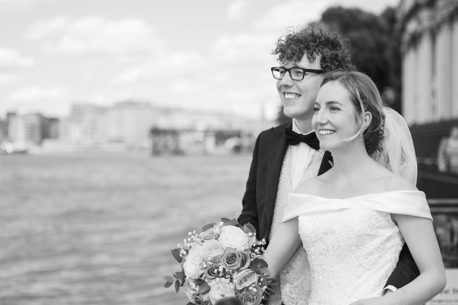 Happy couple standing by the Thames in Greenwich and Trafalgar Tavern, smiling together with the city skyline in the background.