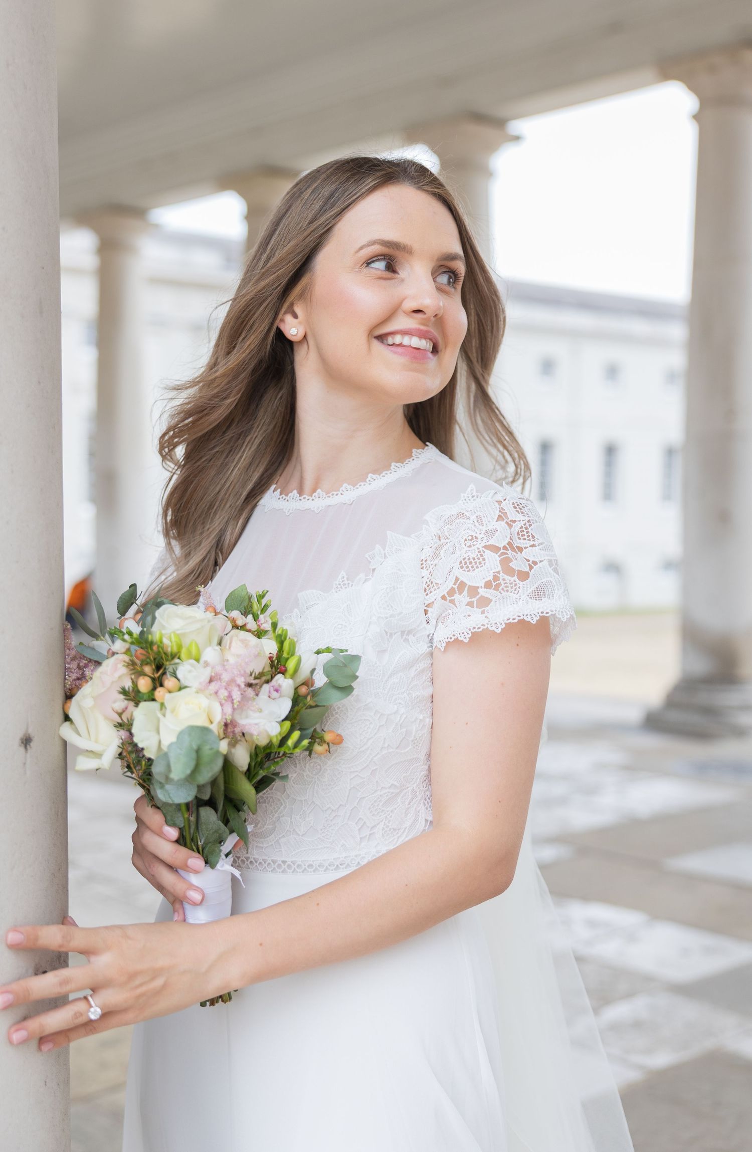 Bride at Queen’s House Column. Smiling bride in lace dress holding her bouquet beside a column at the Queen’s House in Greenwich.