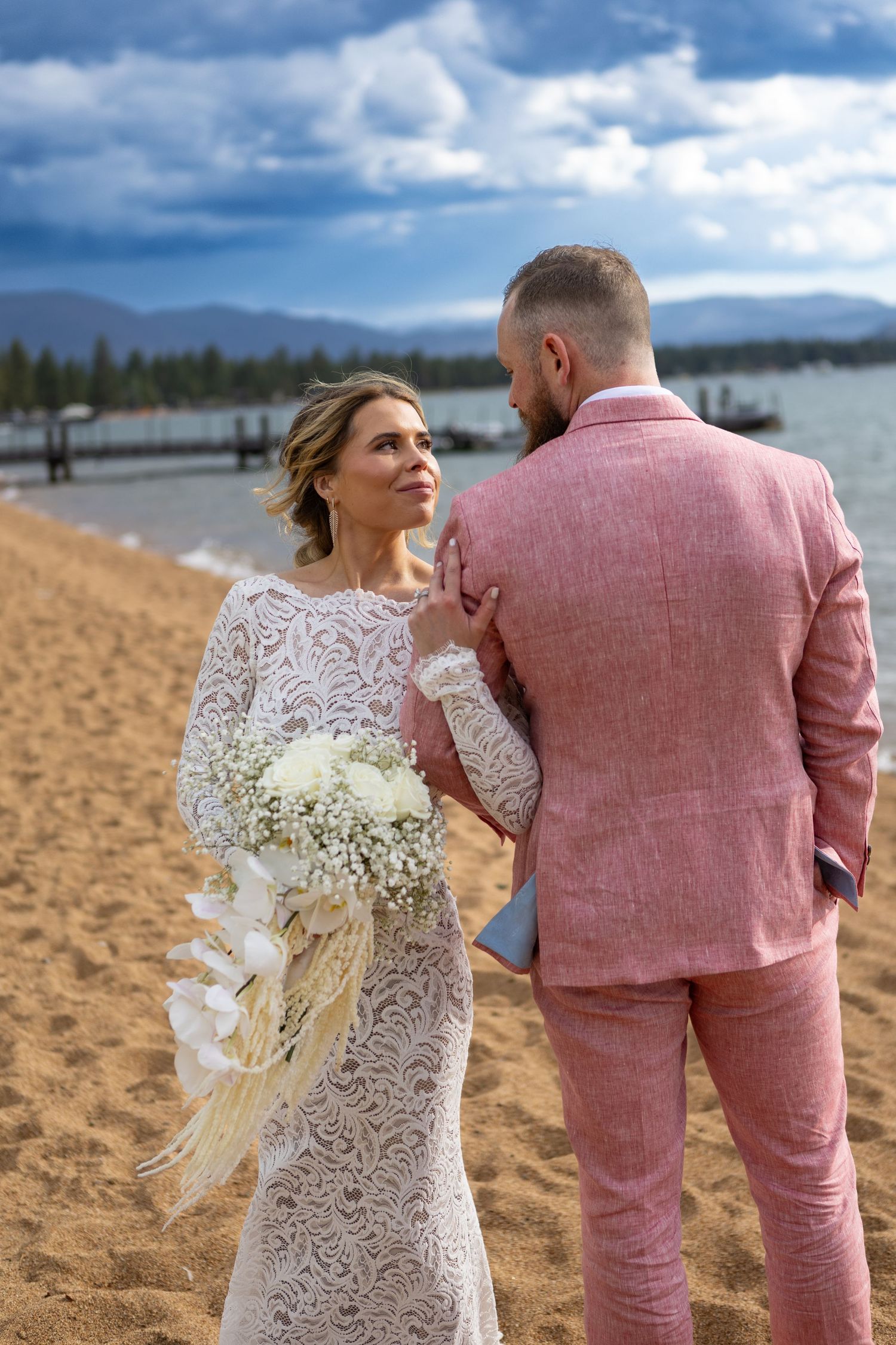 A couple in pink suit and lace dress share an intimate moment on a sandy beach.