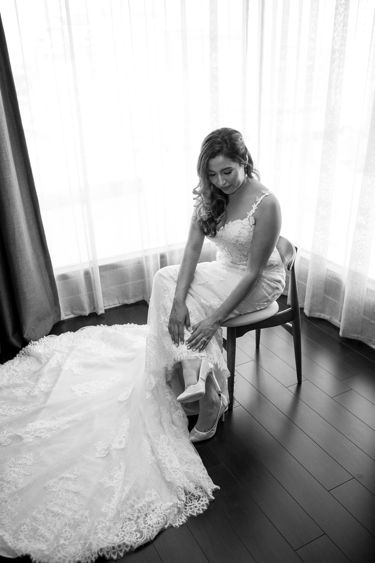 Bride in white lace gown sitting gracefully on dark hardwood floor.