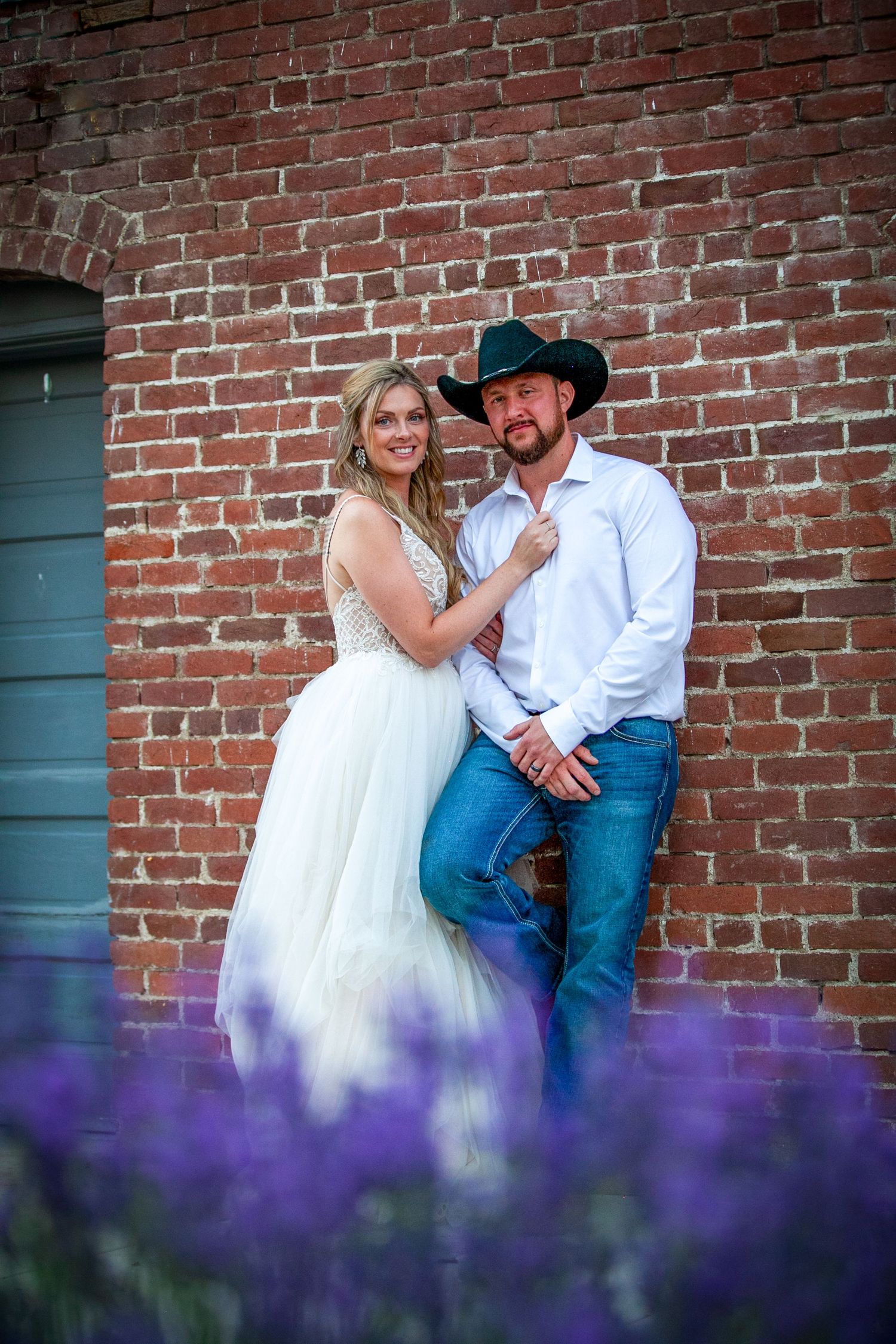 A romantic couple poses against a brick wall with purple flowers in the foreground.