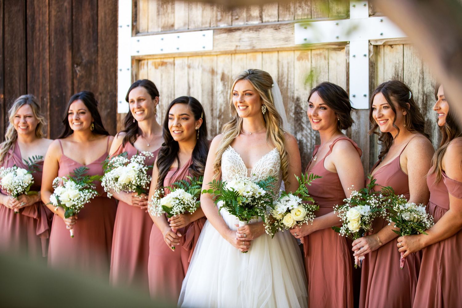 Bridesmaids in dusty rose dresses holding white floral bouquets line up for a group photo.