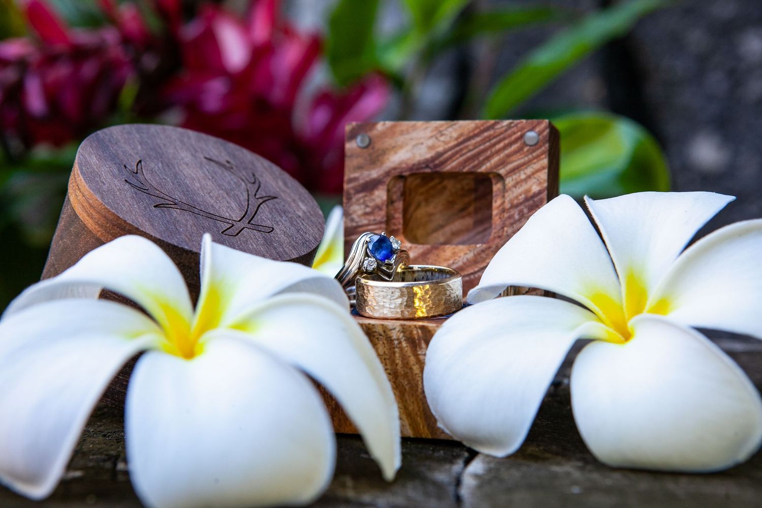Wedding rings nestled in wooden boxes with white plumeria flowers on a stone surface.
