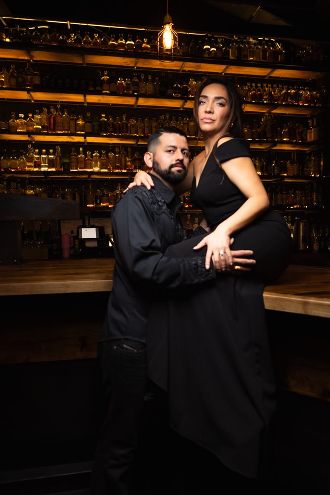 Stylish couple poses at upscale bar with illuminated shelves of bottles in warm lighting.