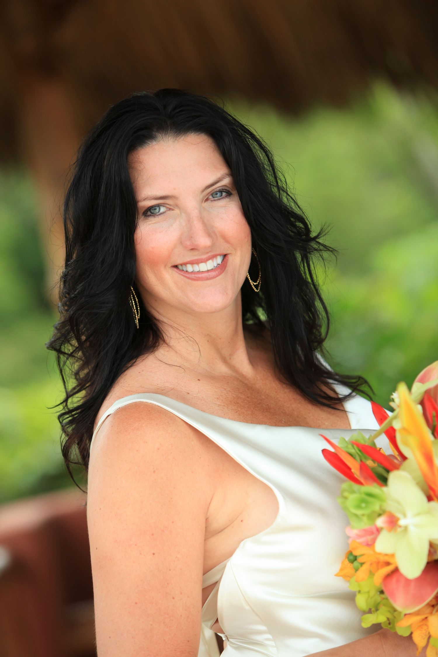 Bride in white satin dress smiles while holding vibrant tropical flowers against a natural background in Costa Rica.