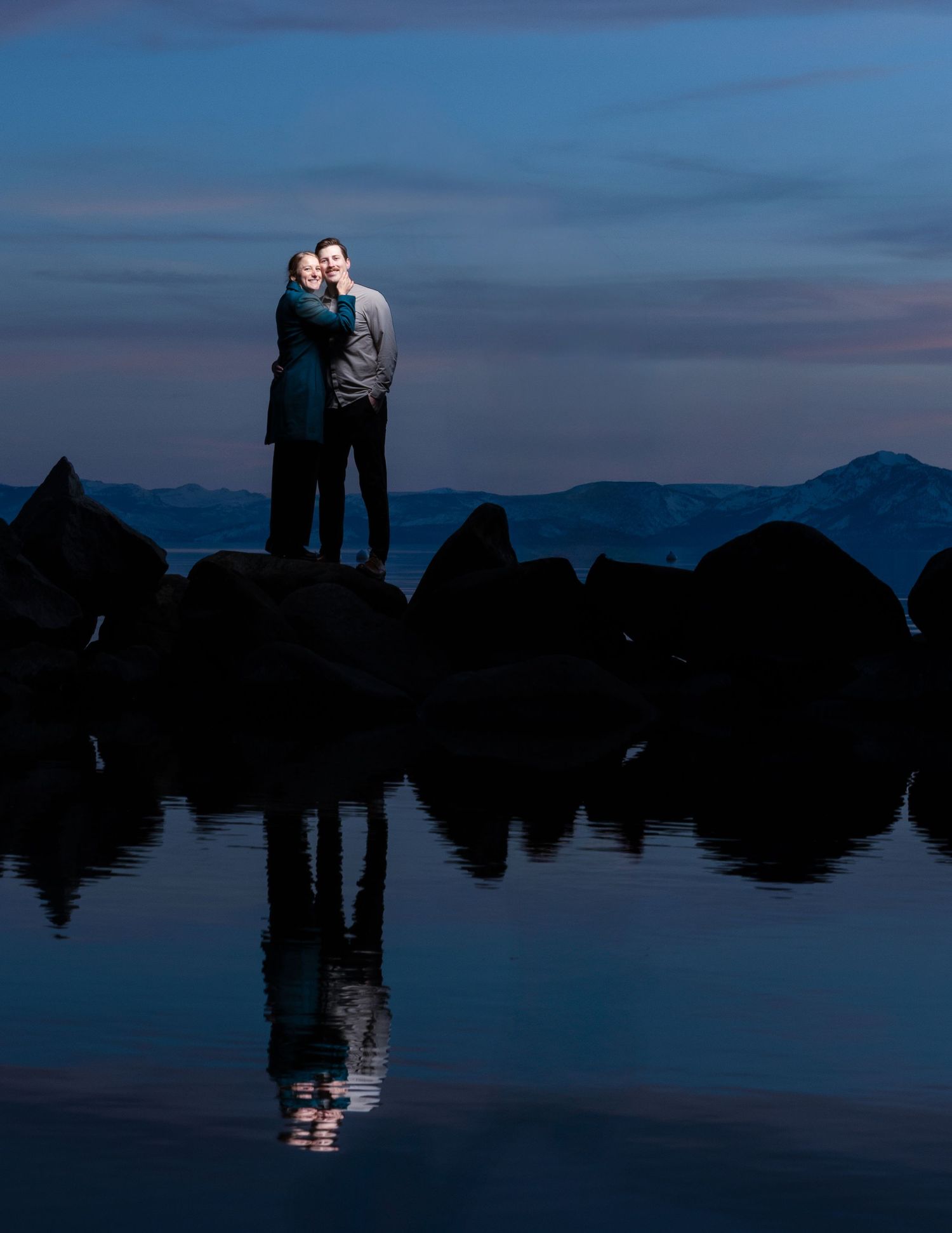 A couple stands silhouetted against a tranquil lake at dusk with mountain reflections creating a serene and dreamy landscape.