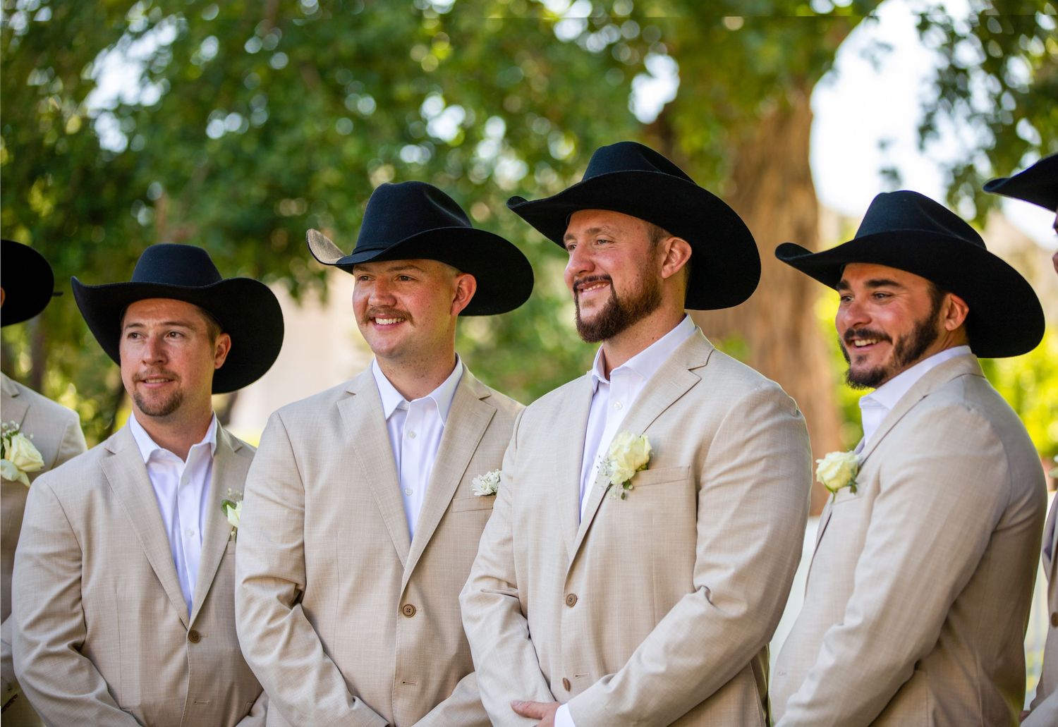 Groomsmen in matching tan suits and black cowboy hats laugh together outdoors.