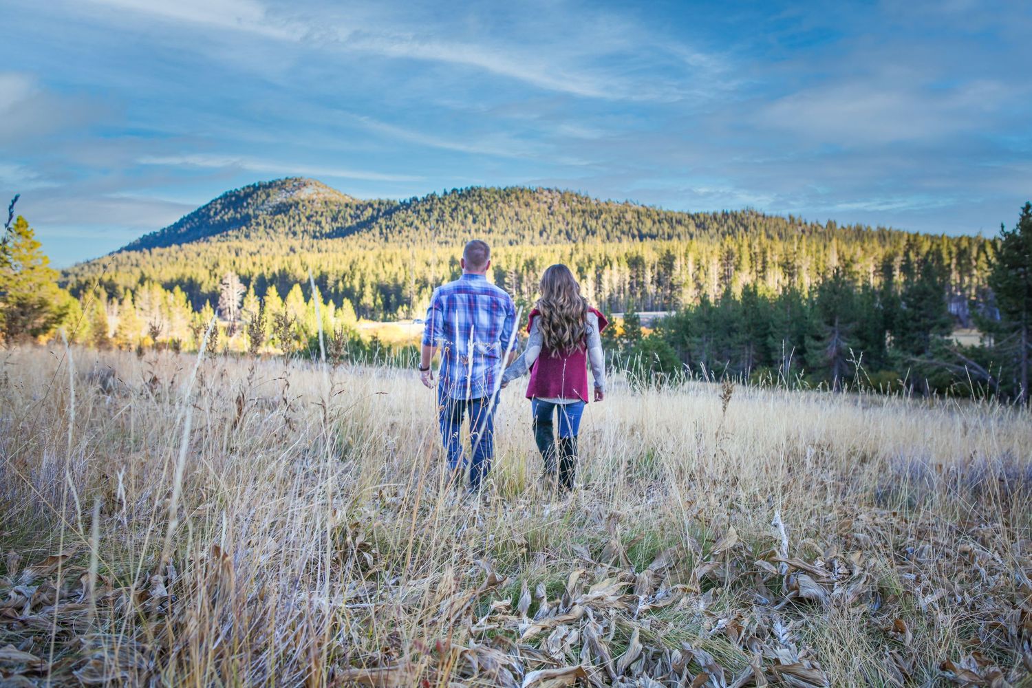 Couple walking through tall grass in autumn field with mountains and evergreen forest in background at sunset.