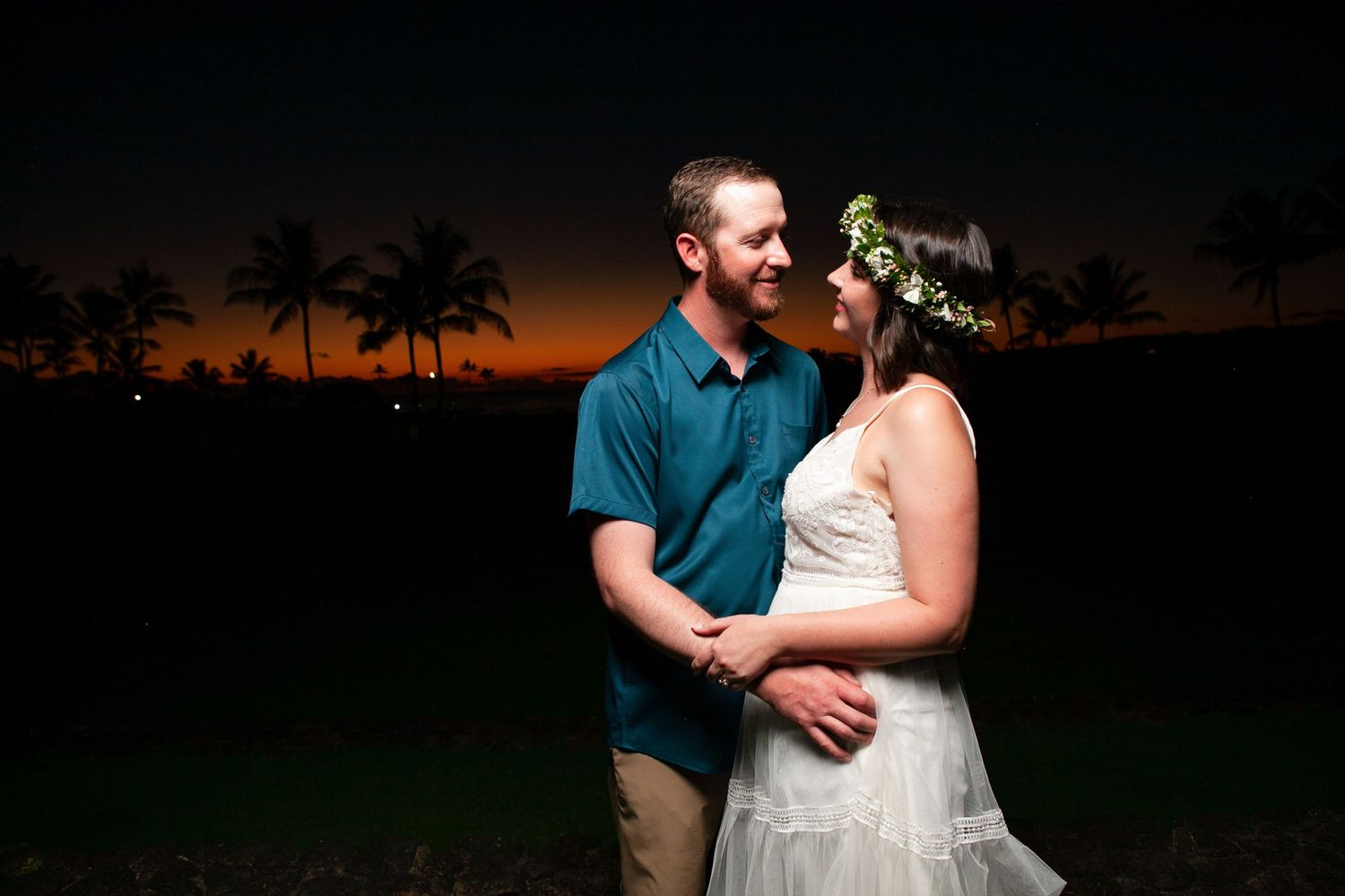 A couple embraces during sunset on a beach with palm trees silhouetted against the orange sky in Kauai.