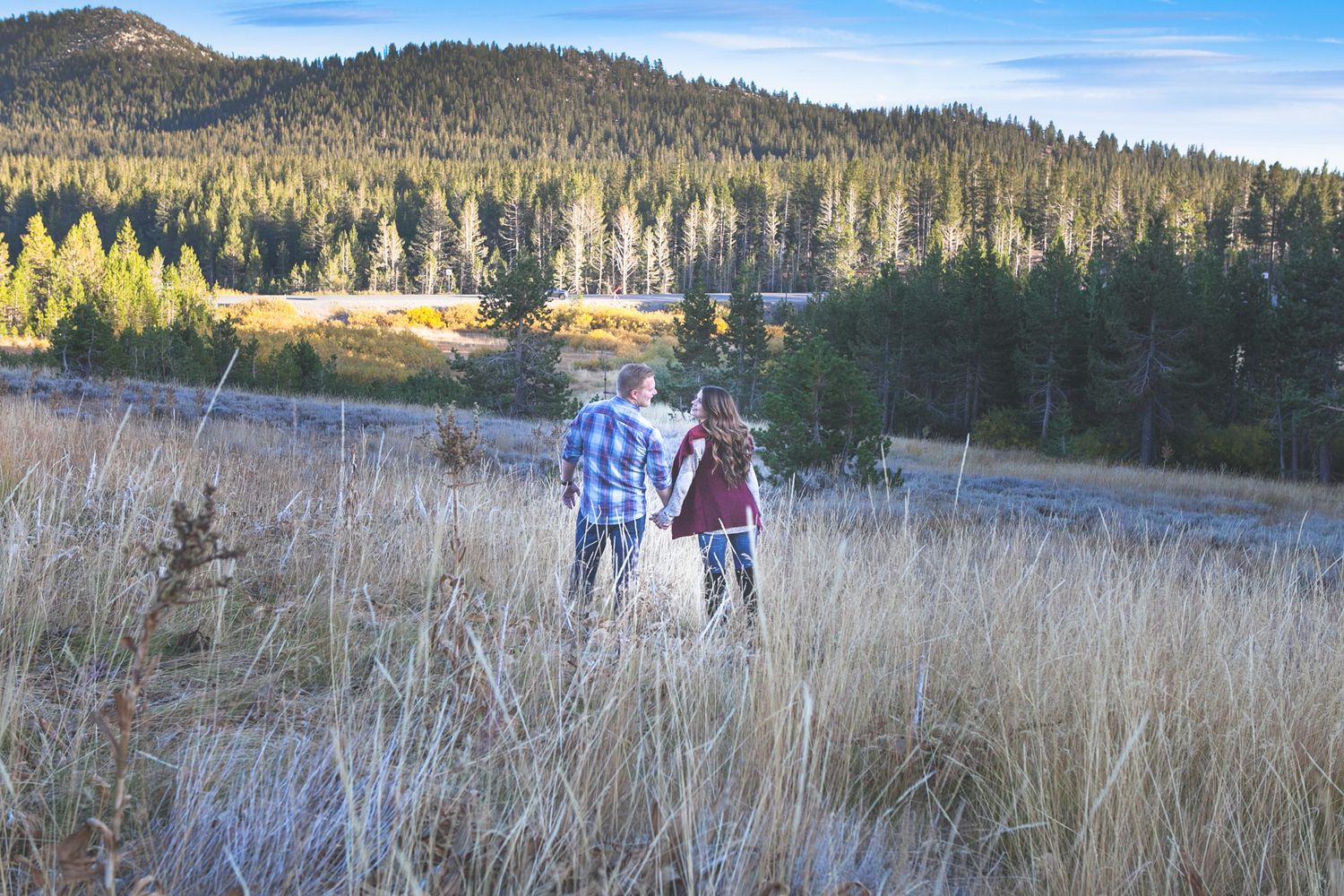 Couple walking through tall grass with mountain landscape in background.