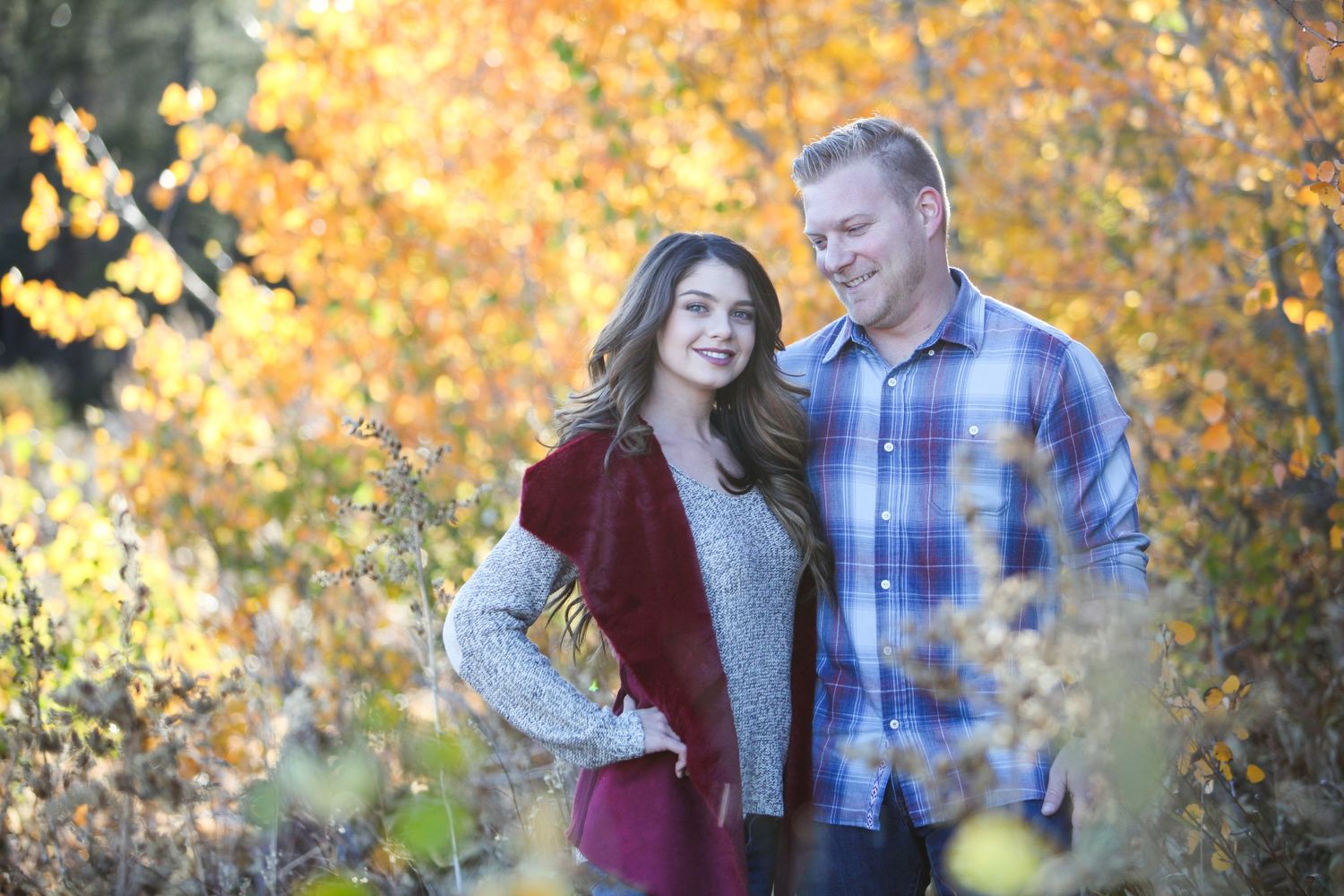 Couple in autumn clothing stands together amid golden fall trees.