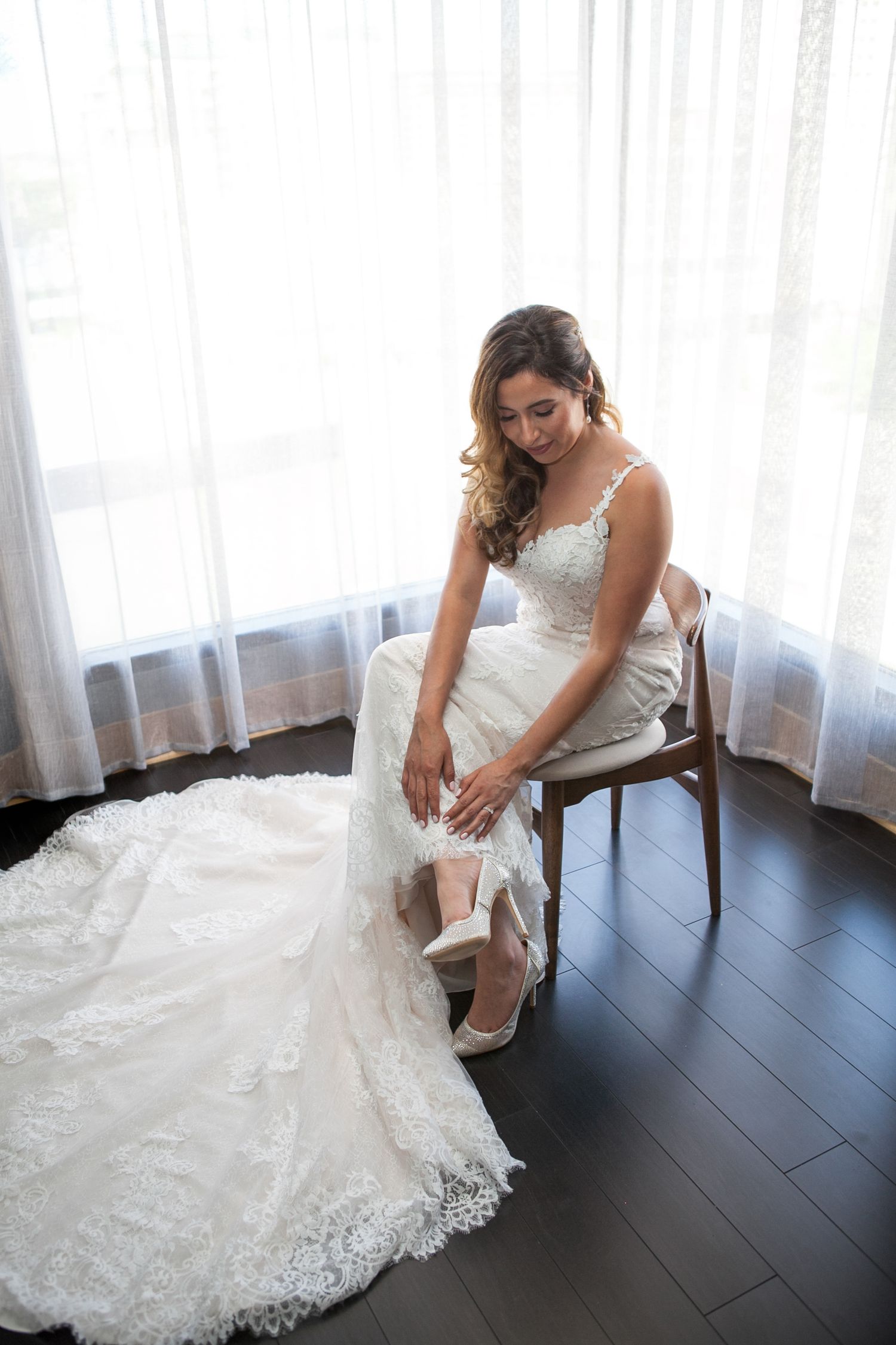 Bride sitting on wooden chair adjusting her shoe with lace dress flowing around her.