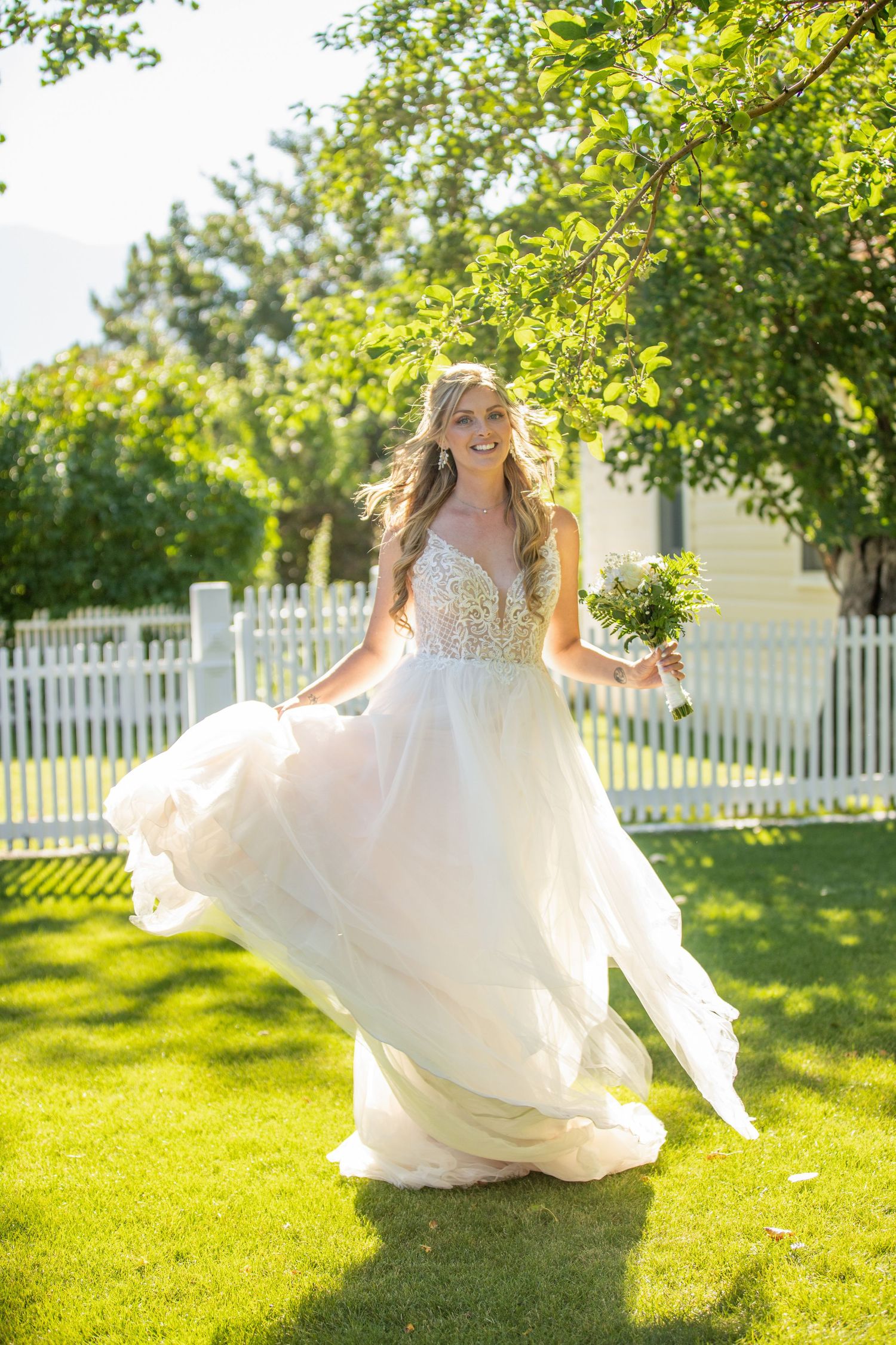 Bride twirls in flowing white wedding dress on sunny lawn with trees in background.