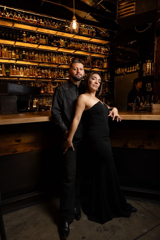Elegant couple in matching black outfits standing in front of well-stocked bar with pendant lighting.