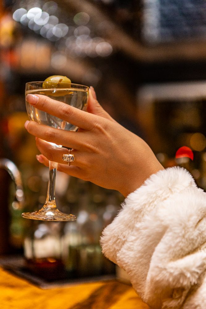 Hand wearing ring holds martini glass with olive garnish in cozy bar setting.