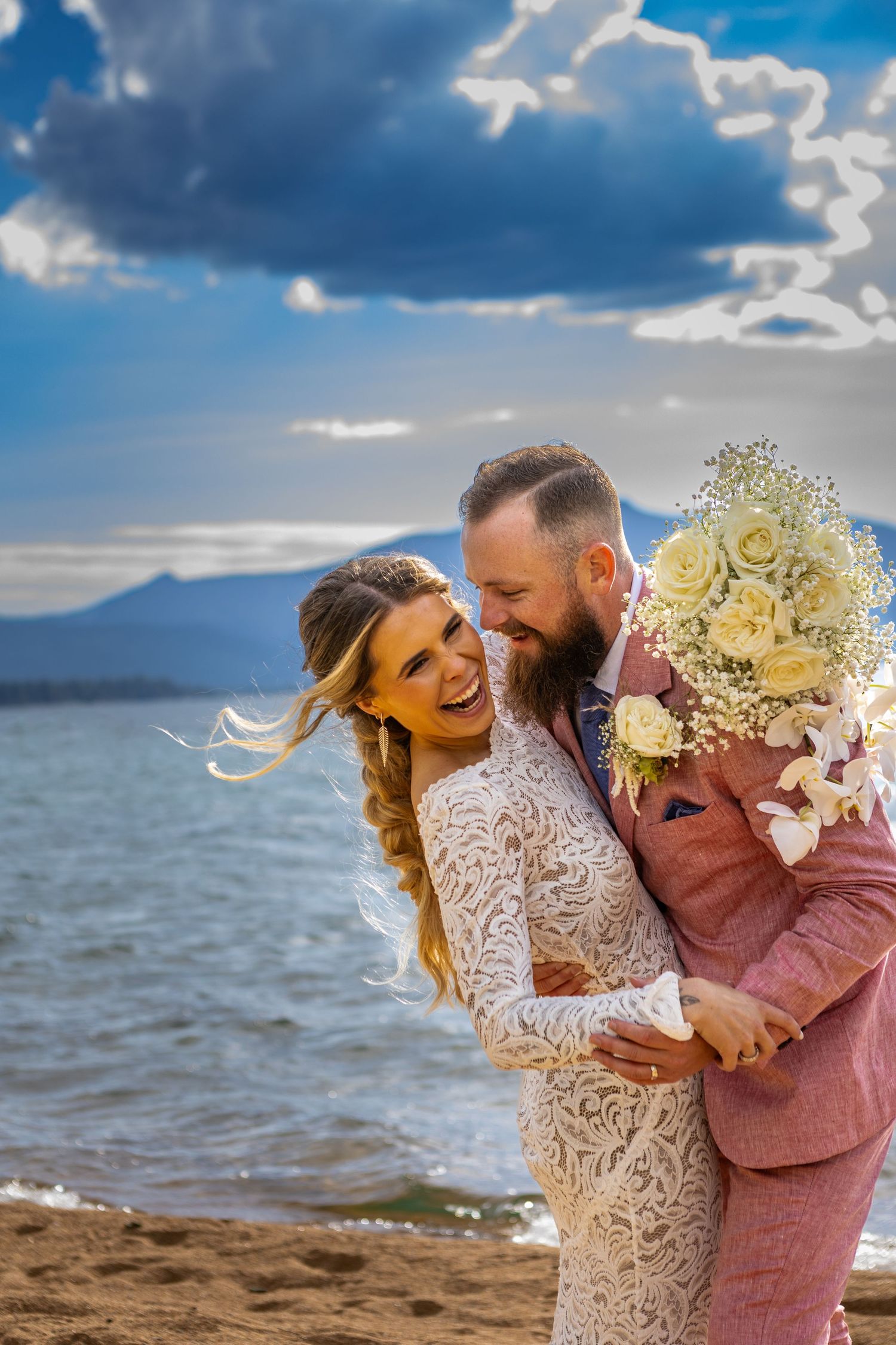 A joyful wedding couple shares a candid moment on the beach with dramatic clouds in the background.