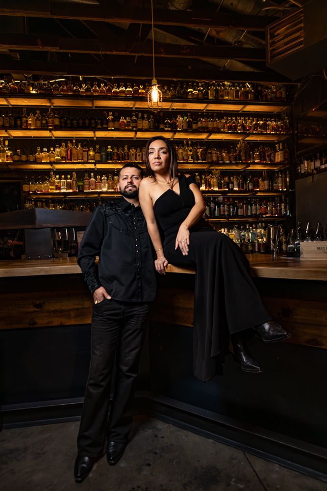 Stylish couple poses at upscale bar with illuminated shelves of bottles in warm lighting.