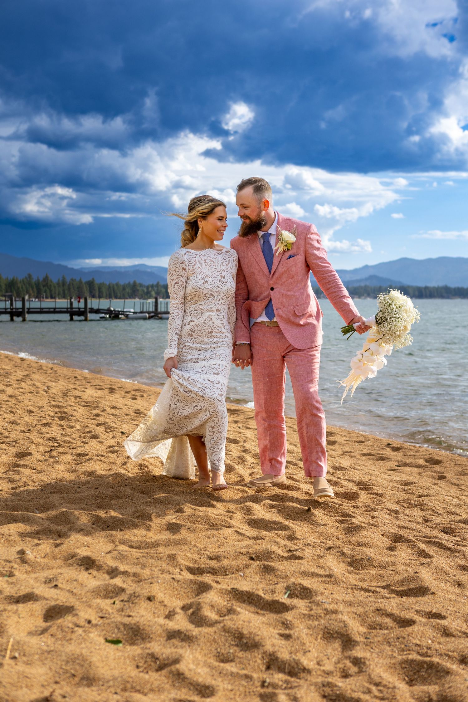A couple in pink suit and lace dress walk along a sandy beach with mountains and pier in the background.