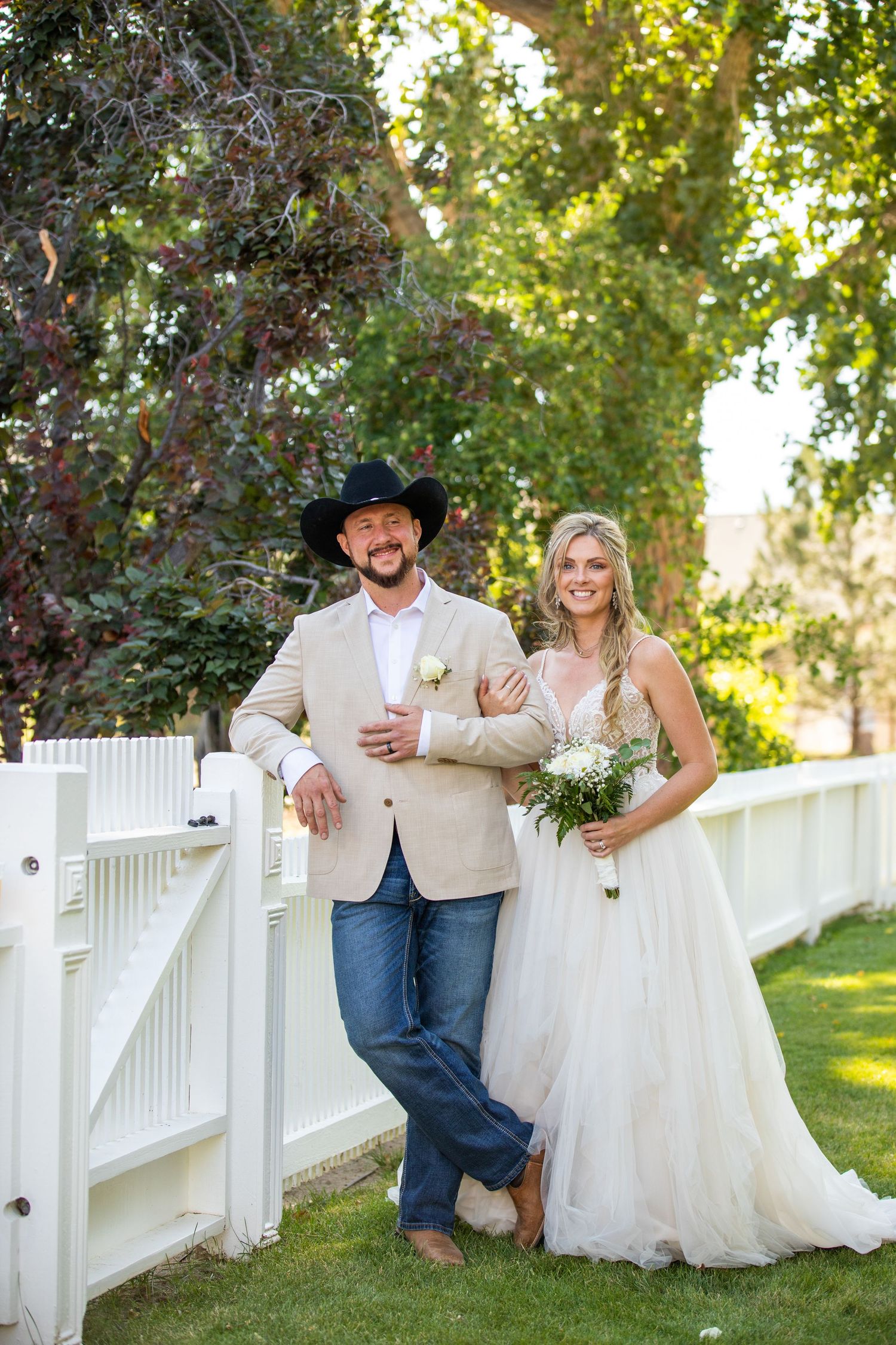 Wedding couple poses playfully along white picket fence in garden setting.