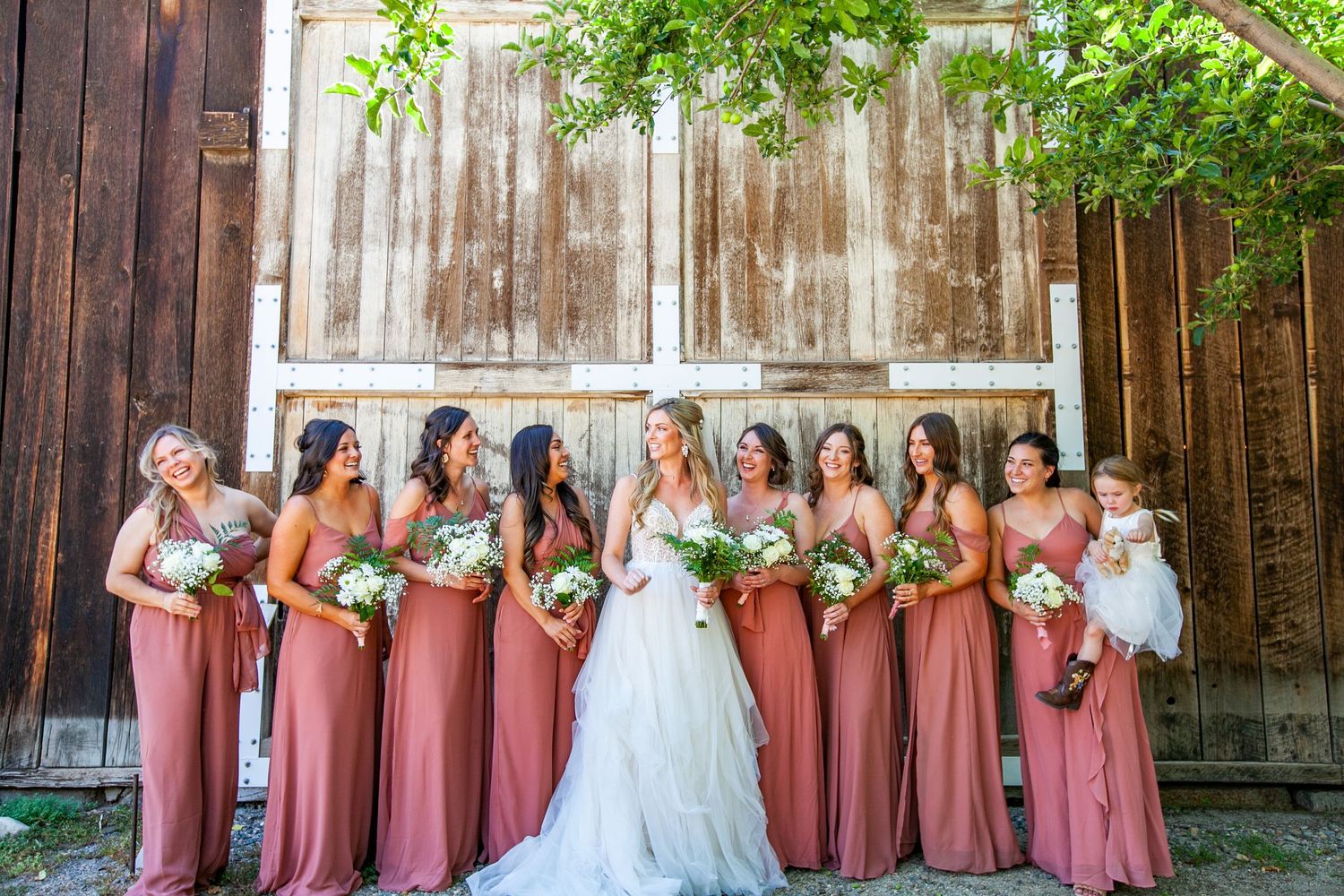 Wedding party in dusty rose dresses poses against rustic barn wall.