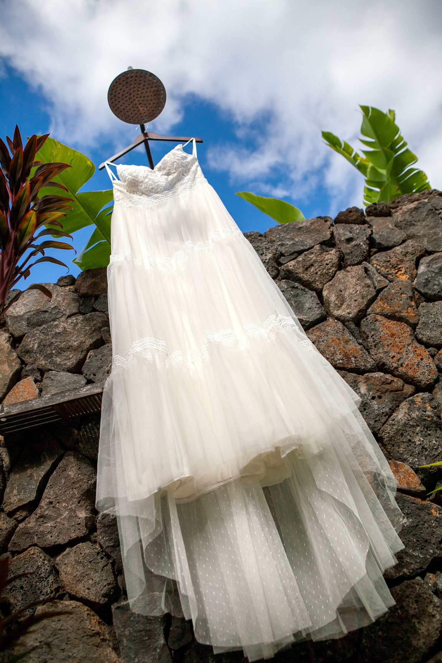 White wedding dress hanging against a stone wall with tropical foliage and blue sky in Kauai.