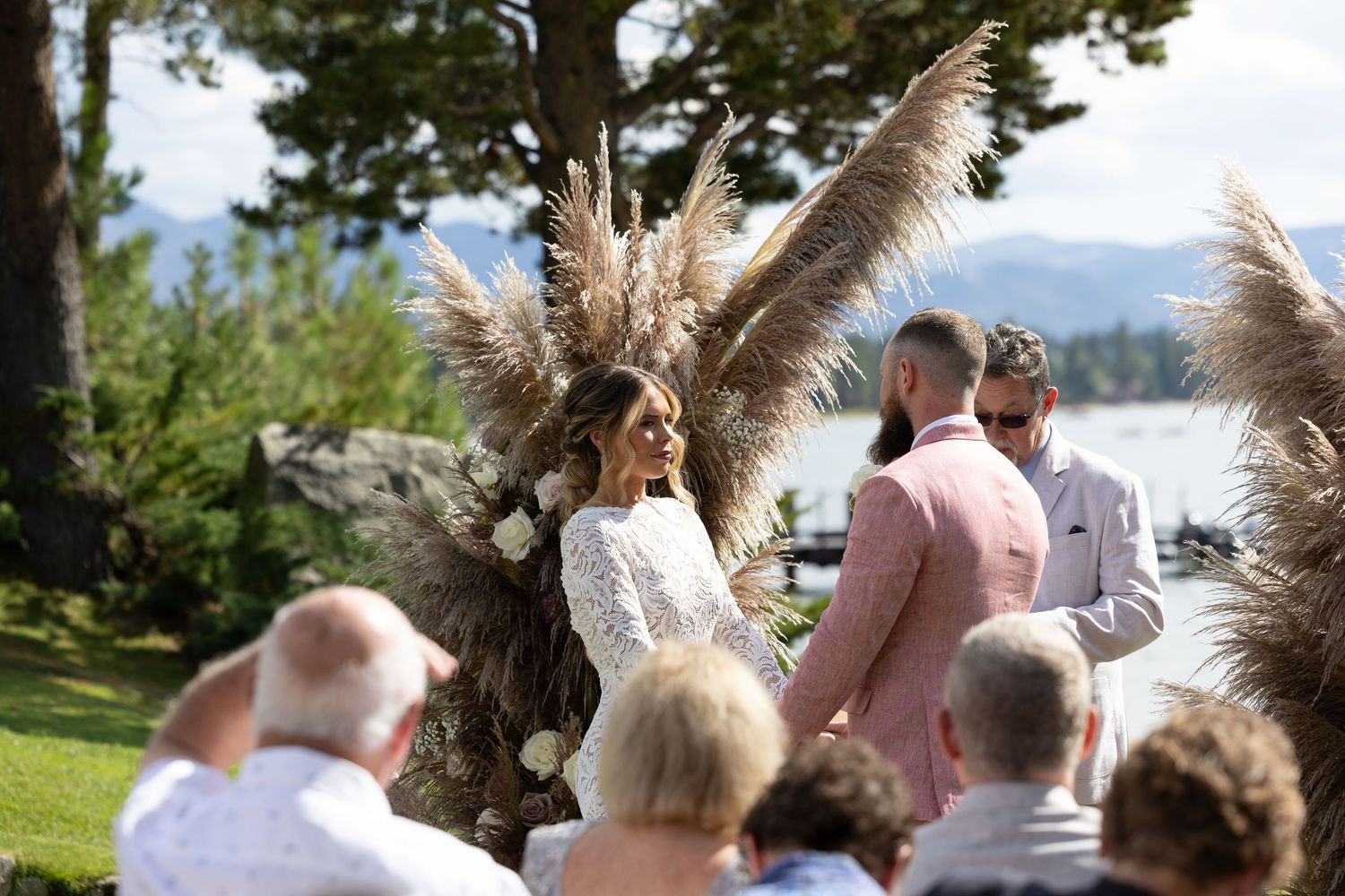 Wedding ceremony guests seated outdoors with pampas grass decorations and lake views.