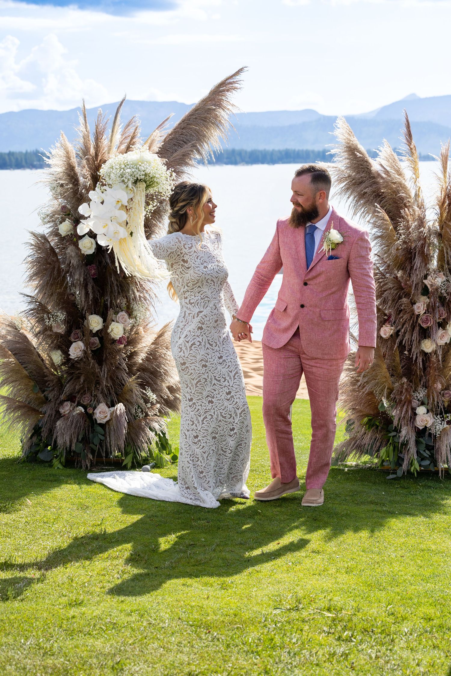 Wedding couple posing by pampas grass arrangements with mountain lake backdrop.