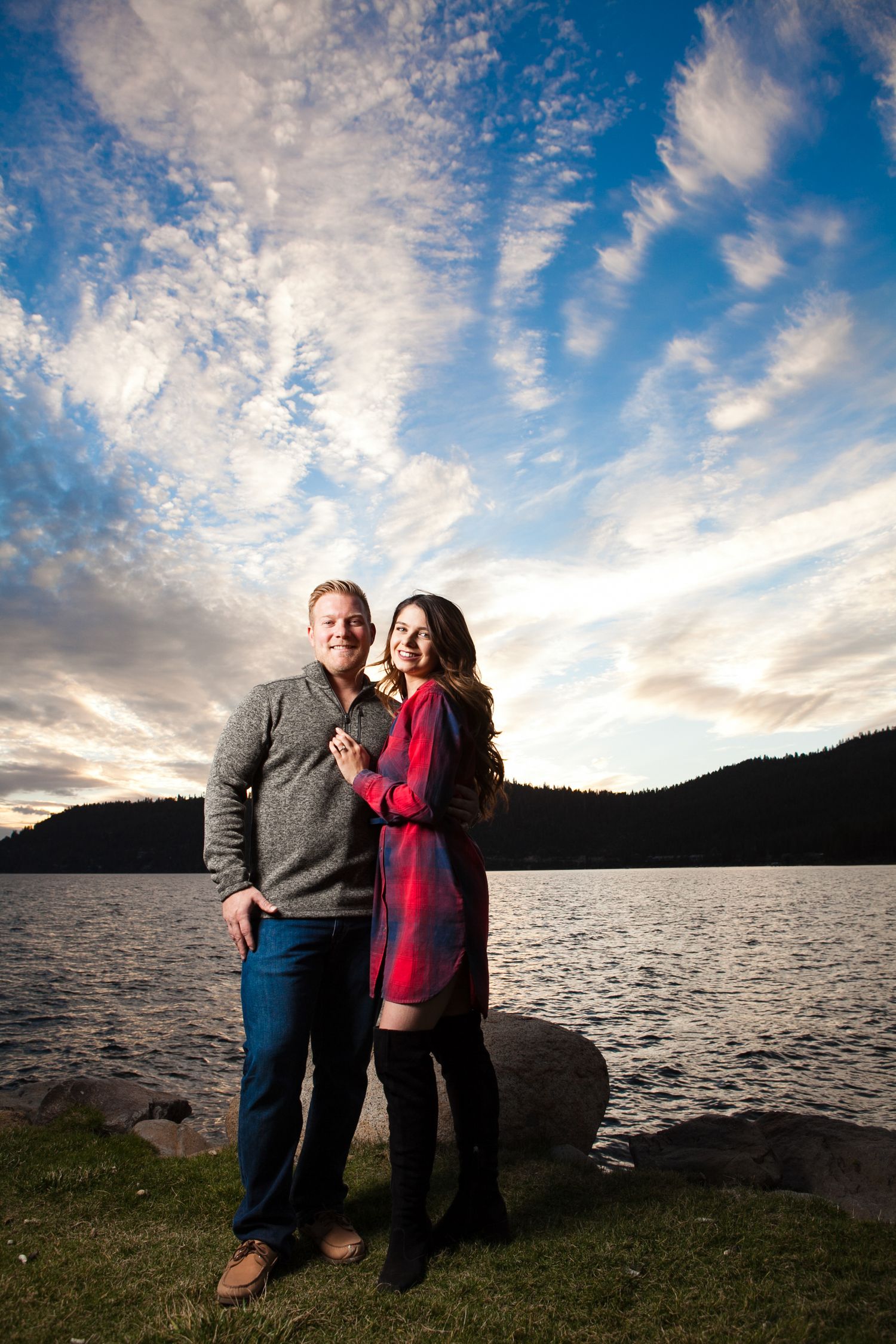 Couple standing together by lake with dramatic clouds and mountain silhouette at dusk in Lake Tahoe.